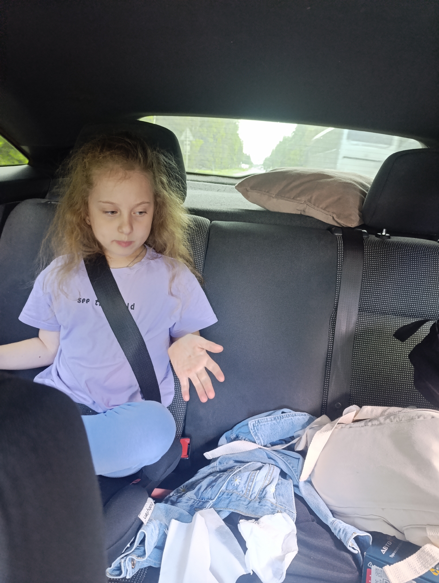 The image shows a young girl with curly hair sitting in the back seat of a car. She is wearing a light purple t-shirt with the words "see the world" and a seatbelt. Her legs are crossed. On the seat next to her are a denim jacket, a white backpack, and a brown pillow. The car is moving, indicated by the blurred trees and a vehicle visible through the back window. The location is Unknown, Unknown.