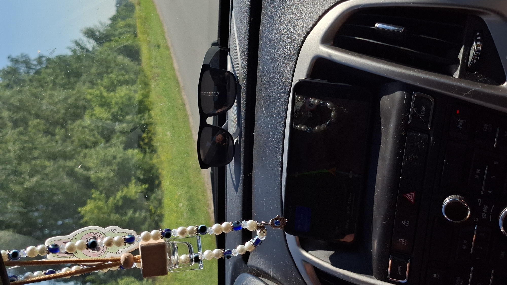 The image shows the interior of a car dashboard on a sunny day, viewed from the passenger seat. On the dashboard, a pair of black sunglasses rests on a textured surface. To the left, a beaded car accessory with a charm hangs from the rearview mirror, partially obscuring the view of the windshield. The accessory is made of white and blue beads with a small wooden cube and a cartoon character. A navigation screen is visible below the sunglasses, displaying a map. The car's central console with various buttons, including a "SPORT" and "TOUR" button, is also visible. Outside the car, through the windshield, a green, treed landscape and a road are visible under a clear blue sky. The overall impression is a snapshot of a car journey in a rural setting.