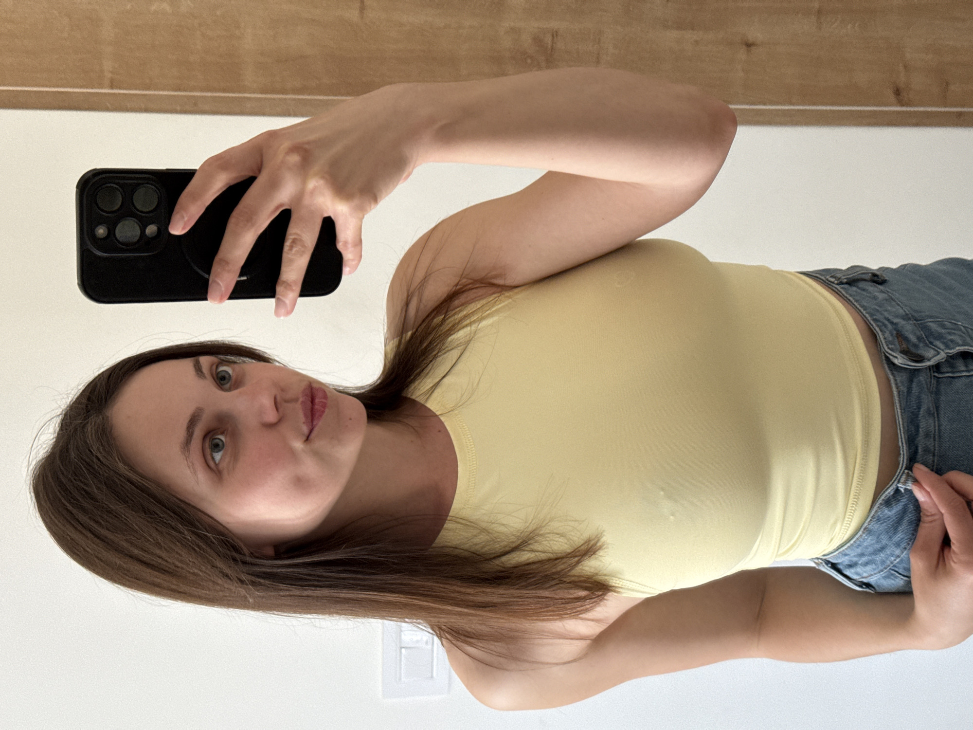 A woman with light brown hair and fair skin is taking a selfie in front of a mirror. She is wearing a yellow tank top and blue jeans. She holds a black smartphone in her left hand. The background includes a plain white wall and a wooden panel, suggesting an interior setting. The location is not discernible from the image, but a location in Belgrade, Serbia is requested. Therefore, one can infer the subject is potentially in Belgrade, Serbia.