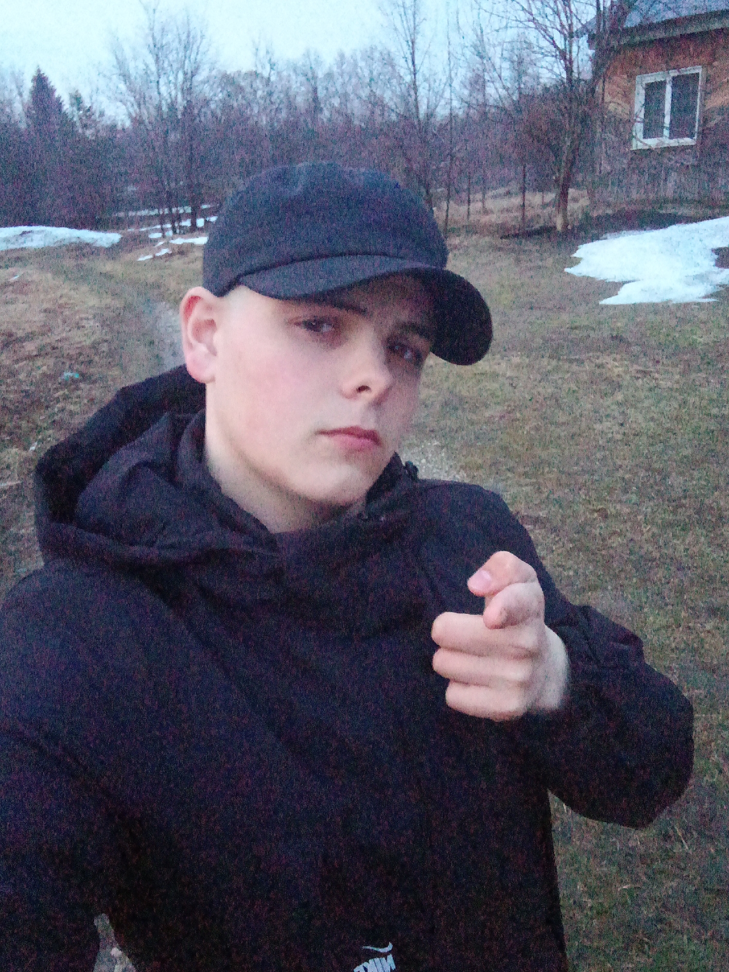 A young, fair-skinned individual, appearing male, is centered in the frame. The person wears a dark-colored baseball cap and a hooded jacket. The jacket hood is pulled up. The individual is facing the camera, with a slight tilt of the head. The right hand is extended towards the camera in a gesture resembling a finger gun, with the index finger and thumb extended.
The background consists of a grassy area, partially covered with remnants of snow. A small wooden structure with a window is visible to the right, indicating a building. Bare trees are scattered in the mid-ground, suggesting late autumn or early spring. The overall setting appears to be a rural or semi-rural environment, in the city of Красный Маяк, Russia. The lighting is diffused, indicating an overcast sky.