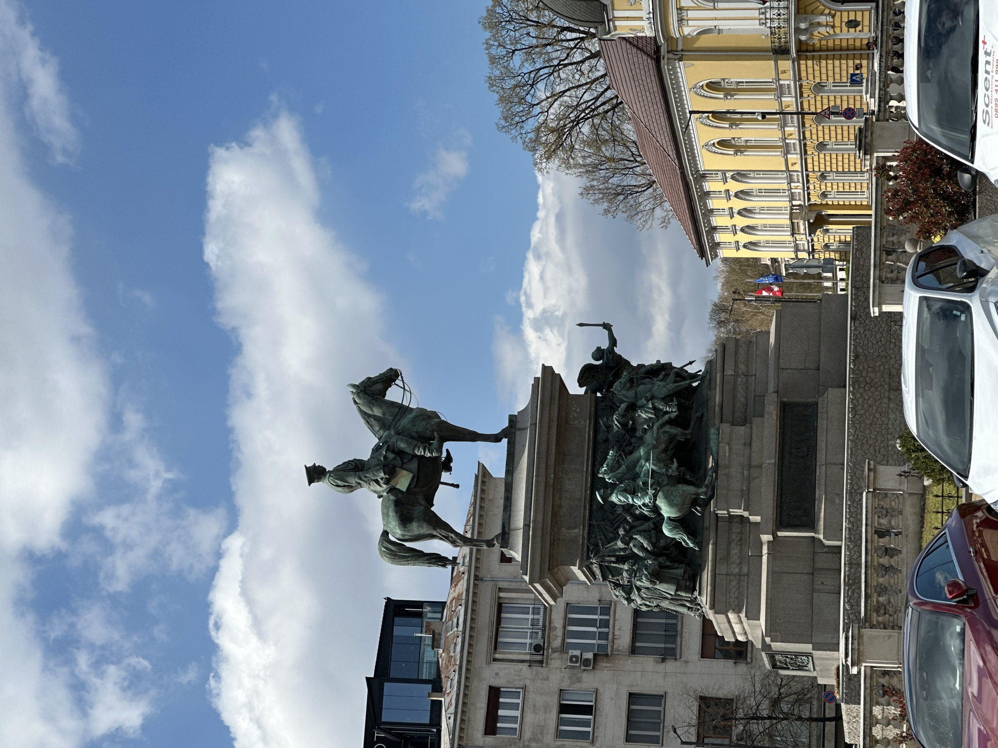 The image shows the Monument to the Tsar Liberator in Sofia, Bulgaria, depicting Tsar Alexander II on horseback. The monument is set against a backdrop of buildings, including a yellow edifice with arched windows and a modern glass-fronted building. The sky is blue with scattered white clouds. Traffic, including cars, is visible on the street adjacent to the monument.