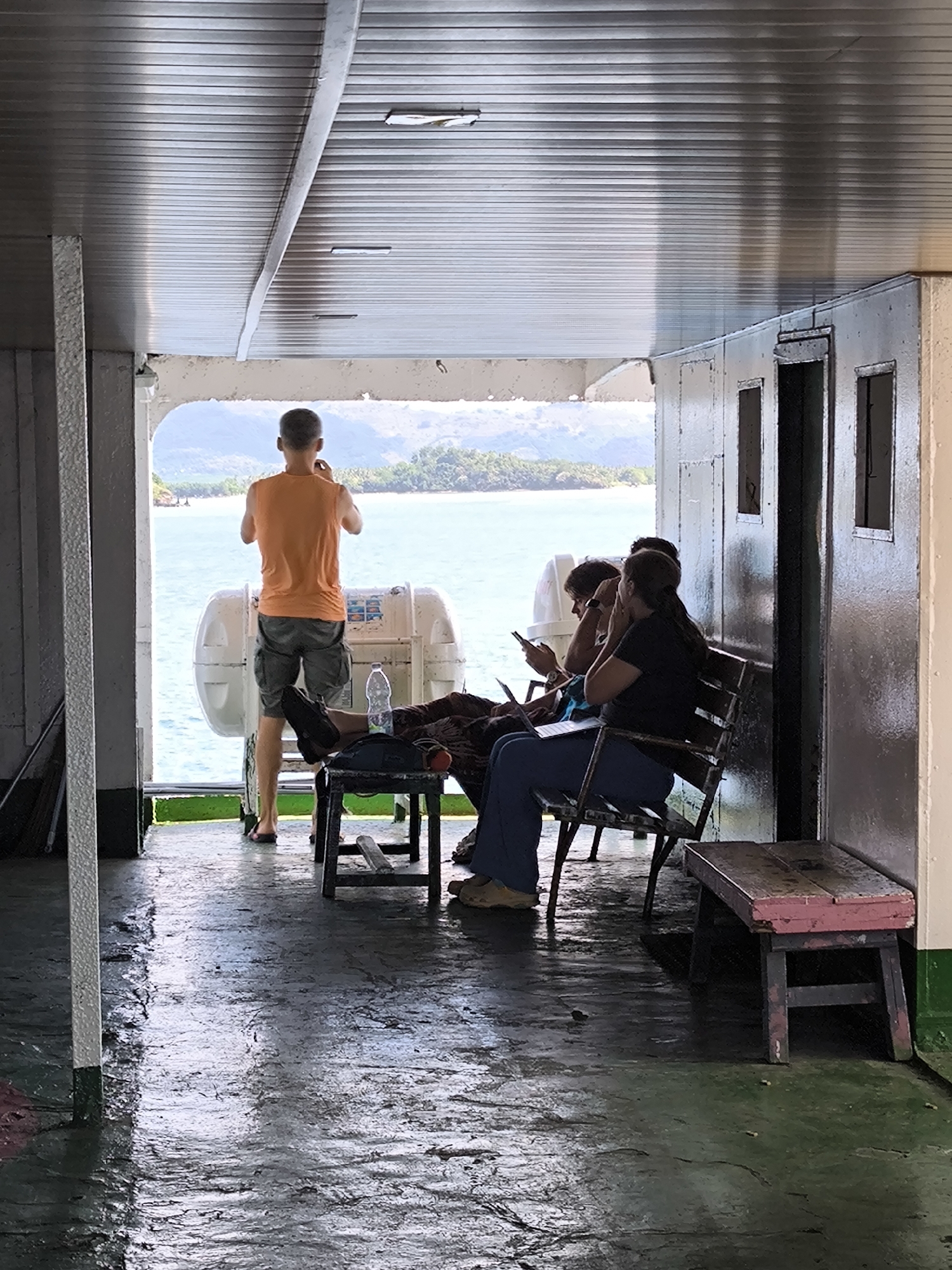 The image captures a candid scene inside a boat or ferry, looking out towards a scenic tropical coastline in Lembar Selatan, Indonesia.
In the foreground, the boat's interior is visible, featuring a dark, wet, and reflective floor that suggests recent cleaning or splashes. A white, vertical column stands on the left. The ceiling is made of reflective, corrugated metal panels.
Three individuals are present. A man in an orange sleeveless shirt and grey shorts stands with his back to the viewer, looking out over the water, holding a phone. He is positioned near a large, white, cylindrical life raft with some illegible red and blue markings. To his right, two other individuals are seated. One woman in a black shirt and blue pants is visible from the side, looking down at a laptop or tablet on her lap. Another person, partially obscured, sits beside her, also looking at a handheld electronic device. They are seated on basic, slatted wooden or metal benches. A small, pink-painted wooden stool is placed on the far right.
Beyond the boat's open stern or side, a calm expanse of light blue-green water stretches towards distant, lush green islands and hills under a bright daytime sky. The overall atmosphere appears relaxed, with passengers either observing the view or engaging with their personal devices during the journey. The light streaming in from the opening suggests it is mid-day. The boat's interior elements, like the corrugated ceiling and metal wall with small openings, give it a utilitarian appearance.
