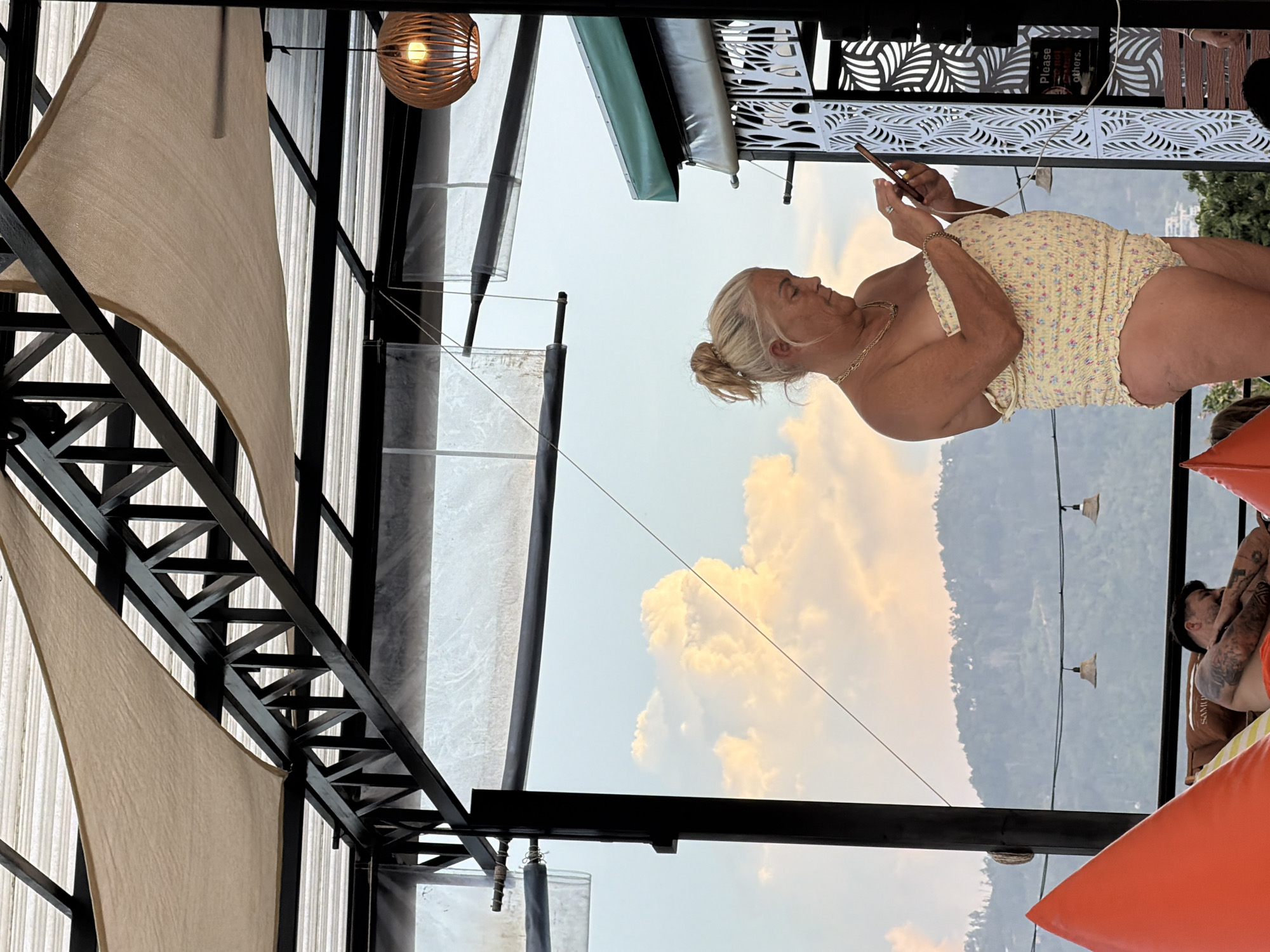 An outdoor scene, likely a resort or restaurant in Ko Samui District, Thailand, captures a woman in a yellow floral-patterned swimsuit looking at her smartphone. She has blonde hair pulled back in a bun and wears a gold necklace. To her left, a man is sleeping on a lounge chair, partially visible behind an orange beanbag. The background features lush green mountains under a sky with scattered clouds tinged with orange hues, suggesting either dawn or dusk. A wooden-slatted pendant light hangs from the ceiling structure, which is supported by dark metal beams and covered with a light-colored fabric awning.