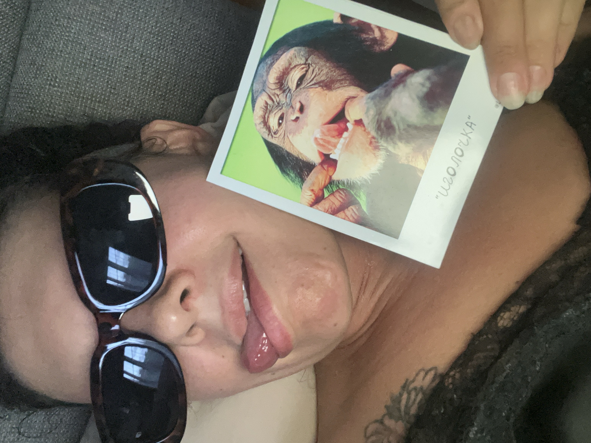 A woman lies down wearing sunglasses, with her tongue sticking out. She is holding a photo of a chimpanzee eating. The photo is labeled "Иголочка" in Cyrillic script. There are no indications that the image was taken in North Kuta, Indonesia.