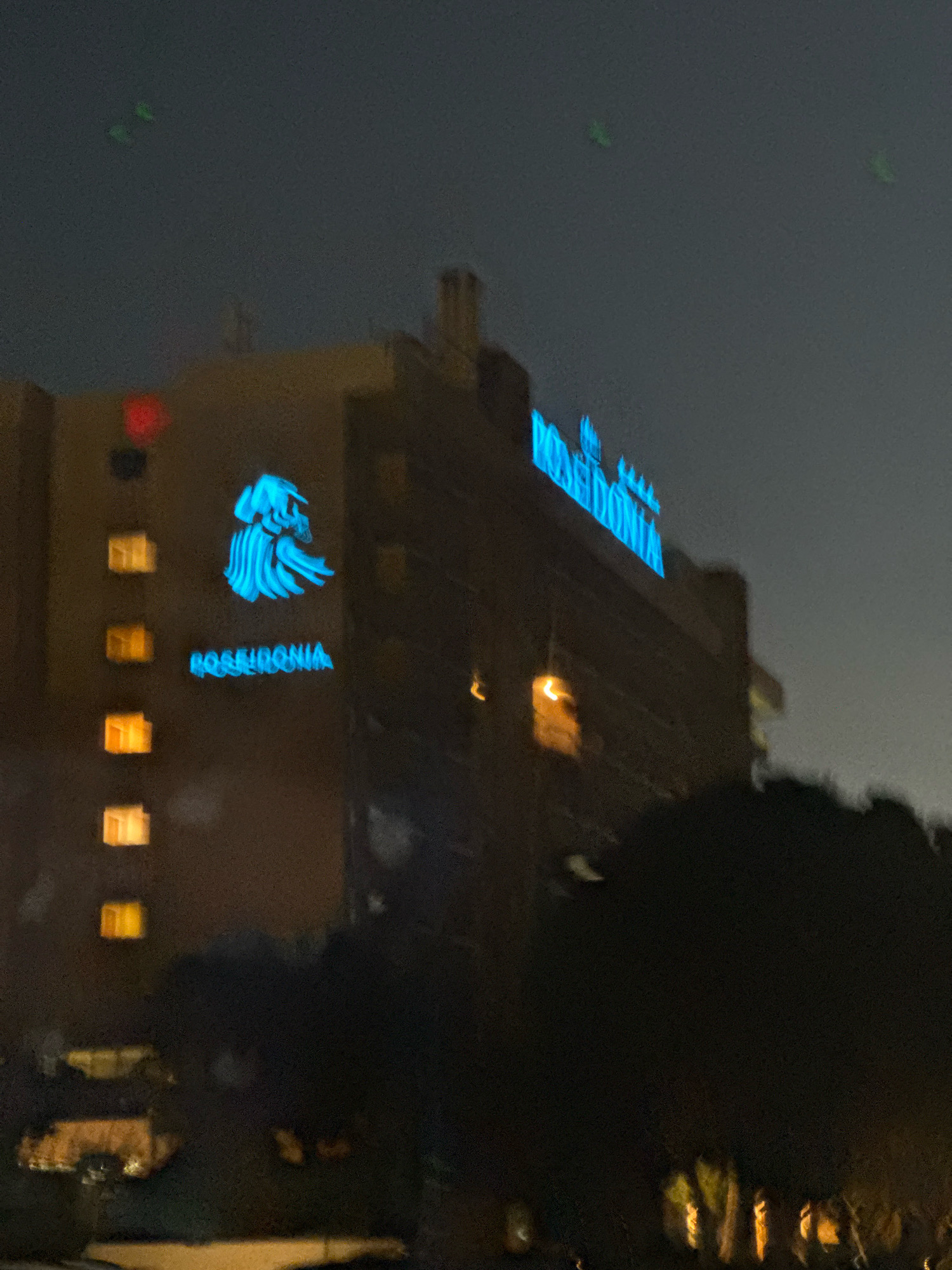 The image depicts a nighttime scene of the Poseidonia Hotel in the Agios Tychona Community, Cyprus. The hotel is a multi-story building with illuminated windows. A prominent blue neon sign on the hotel displays the name "POSEIDONIA" along with a logo featuring a stylized head. On the left side of the building, a blue neon logo of a head is also visible. There are several green lights in the dark sky above the hotel. The foreground is dominated by dark silhouettes of trees. The overall impression is of a hotel at night, likely in a coastal area given its name and location in Cyprus.