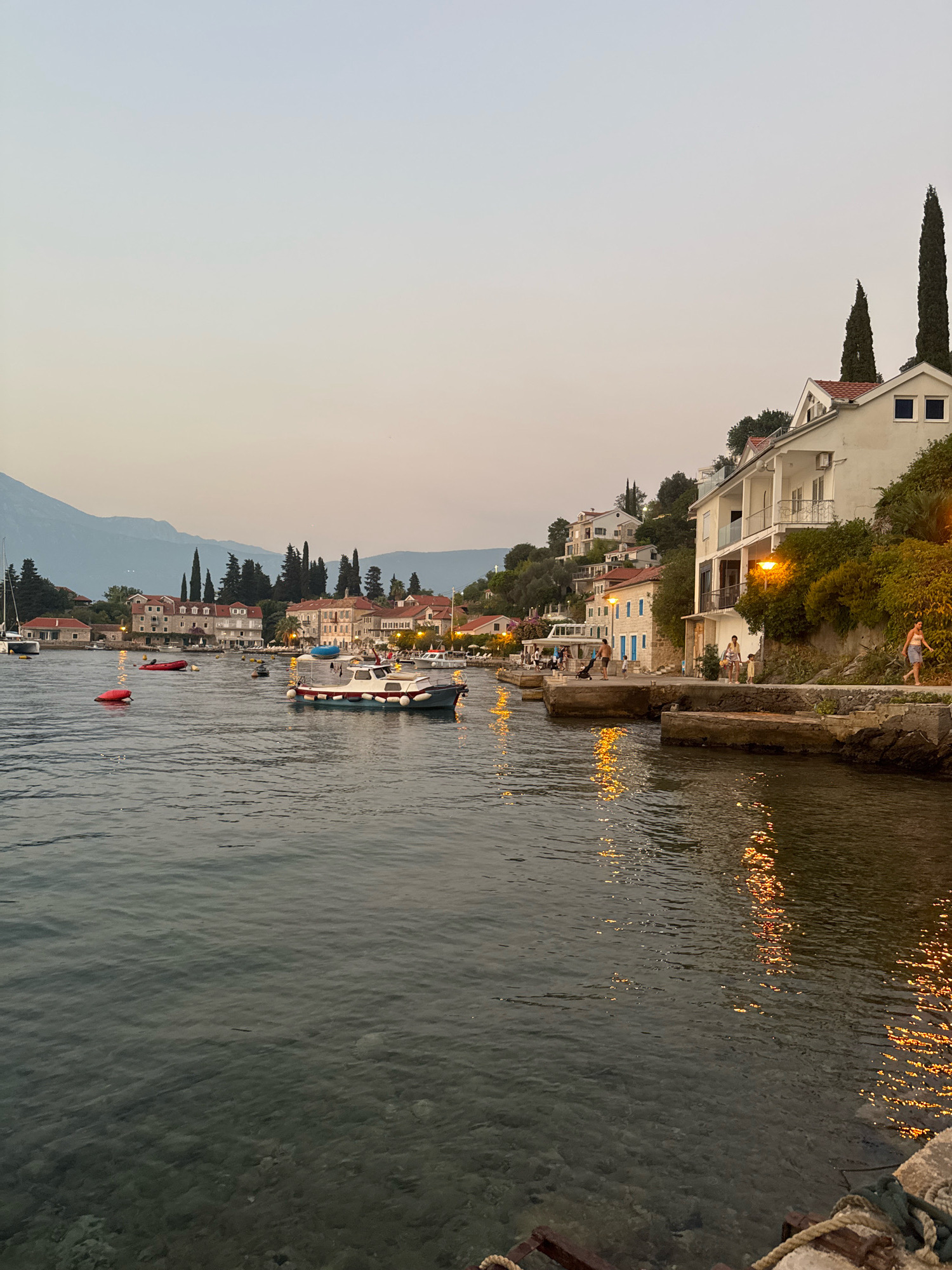 The image depicts a waterfront scene in a coastal town, likely at dusk or early evening. The water in the foreground is dark and reflective, with the lights from buildings on the shore creating shimmering patterns. Several boats are moored or moving in the bay, including a white and blue motorboat closer to the shore and a red inflatable boat further out. Along the waterfront, there are multi-story buildings with red-tiled roofs, some of which are lit from within. A narrow walkway or road runs along the front of these buildings, and several people are visible strolling or standing. One woman in a light-colored dress is walking along the paved walkway, and a few other figures are present further down the street. A gentle slope rises behind the buildings, covered in lush green vegetation and dotted with more houses and tall, slender cypress trees. In the background, hazy mountains rise against a pale sky, suggesting a scenic, possibly Mediterranean, location. The overall atmosphere is tranquil and picturesque.