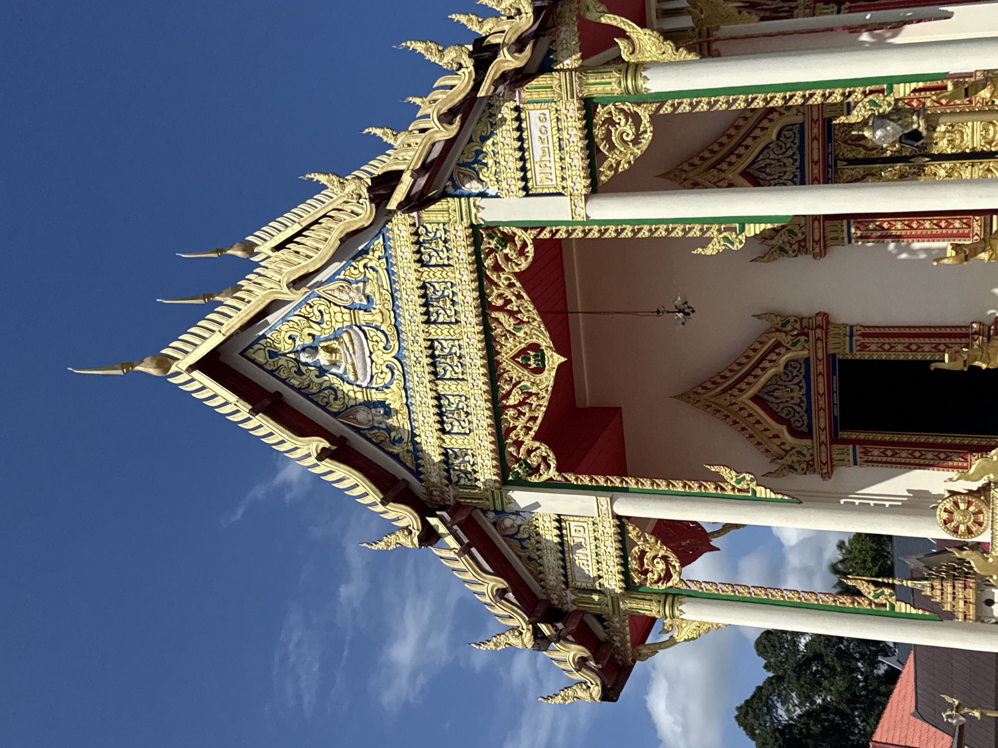 The image shows an ornate Buddhist temple in Baan Taling Ngam, Thailand, viewed from a low angle. The structure features a multi-tiered roof with intricate gold decorations, red accents, and blue and white details. White pillars with green and gold trim support the roof overhang. The sky is a clear blue with scattered white clouds. Trees and parts of other buildings are visible in the background.