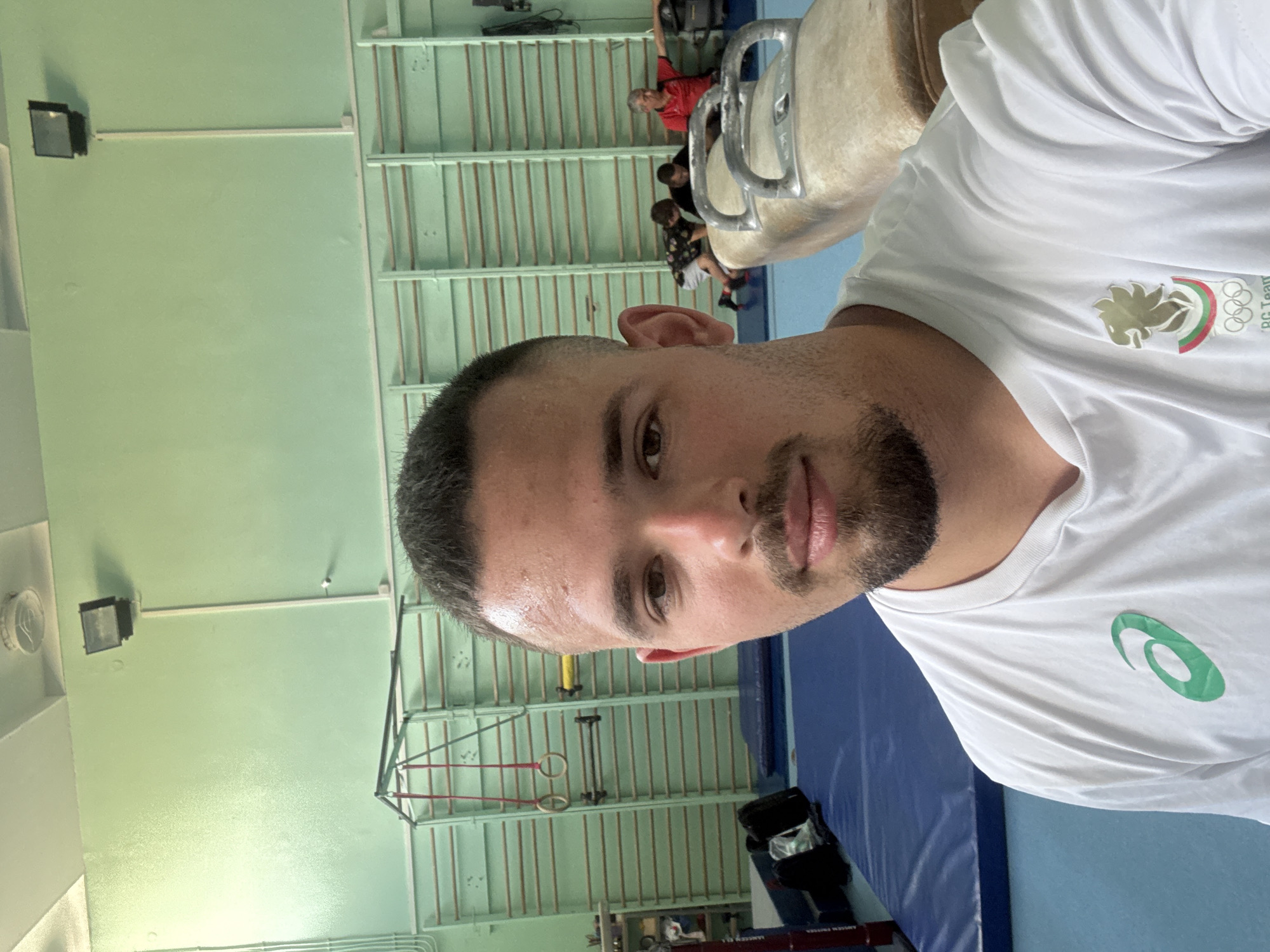 A man takes a close-up selfie from the chest up within a gymnastics training facility in Blagoevgrad, Bulgaria. He has short dark hair and a well-kept beard, looking directly at the camera with a neutral expression. Sweat glistens on his forehead and face, suggesting recent physical activity.
He is wearing a white short-sleeved t-shirt. On the left side of his chest, an emblem features a golden, stylized bird or phoenix-like figure, accompanied by a small tricolor stripe resembling the Bulgarian flag (white, green, red), the text "BG Team," and the Olympic rings. On the right side of his chest, a prominent green "Asics" logo is displayed.
The background shows an indoor gymnasium. A large blue mat covers part of the floor. To the man's right, a pommel horse is partially visible. Further back, various gymnastics equipment, such as structures for parallel bars or rings, and wall bars, line a light green wall. Blurred figures of other people are dimly seen in the background. Fluorescent lights illuminate the interior space, consistent with a training environment.