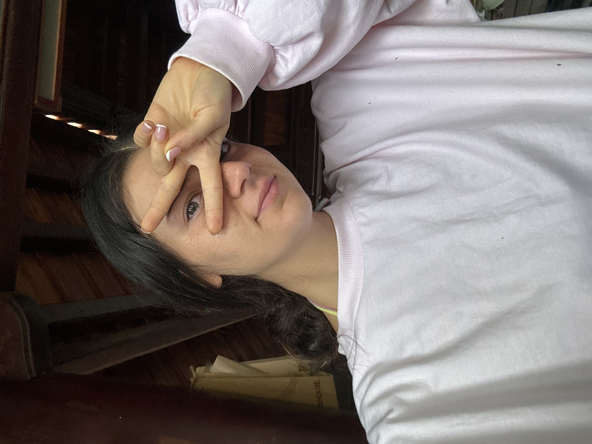 A young woman with dark hair and fair skin is pictured indoors, holding her hand up to her face, with fingers spread in a gesture that partially obscures her left eye. She is wearing a light pink, long-sleeved sweatshirt with cuffed sleeves. The background appears to be a dimly lit wooden structure, possibly stairs. At the bottom of the image is a stack of books. The location of Mũi Né, Vietnam, is not verifiable from the image alone.