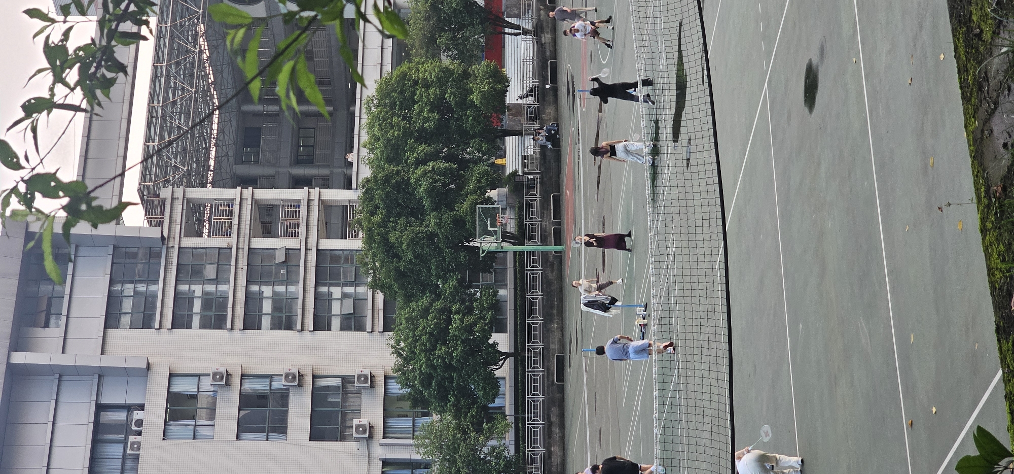 Yuzhong District, China. Exterior view of a public tennis court with people playing. The court is surrounded by a fence and trees. In the background, there is a building. Several people are standing on the court.