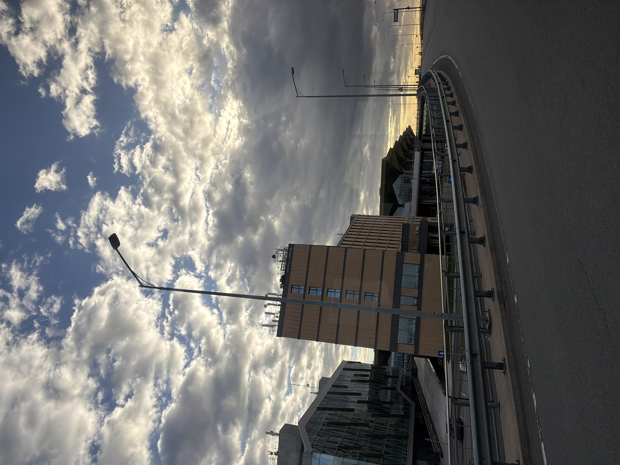 The image depicts a cityscape in Saint Petersburg, Russia. It features a modern architecture building complex with a tan and brown office building and a lower glass-covered structure. Streetlights are visible along the roadside. The sky is cloudy, with blue patches interspersed with gray and white formations. The foreground includes a section of elevated highway, with a gray guardrail running alongside. The overall perspective is from a low angle, looking upwards. The "Hotel Отель" sign is visible on the building. The scene appears to be captured near Pulkovo Airport based on the architecture.