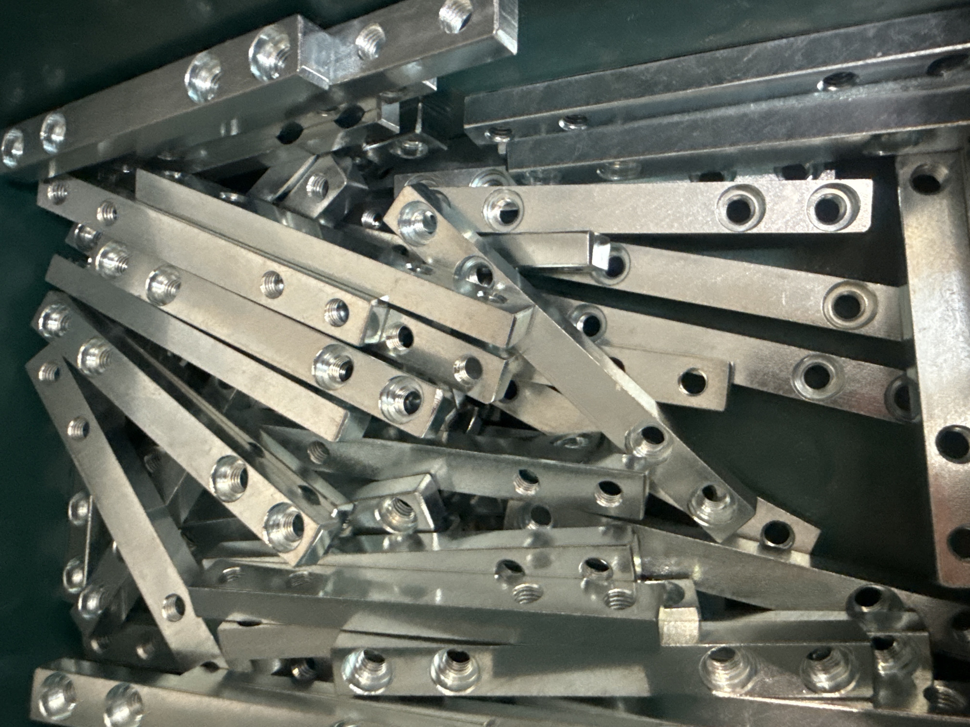 A pile of shiny, metallic, rectangular components are tightly packed together in a dark green container. These components appear to be manufactured metal parts, each featuring multiple circular holes, some of which are threaded. The lighting casts strong reflections off the metallic surfaces, creating bright highlights and deep shadows, obscuring some of the finer details of the pile. The composition is a close-up, overhead shot. This image was taken in Fontevivo, Italy.