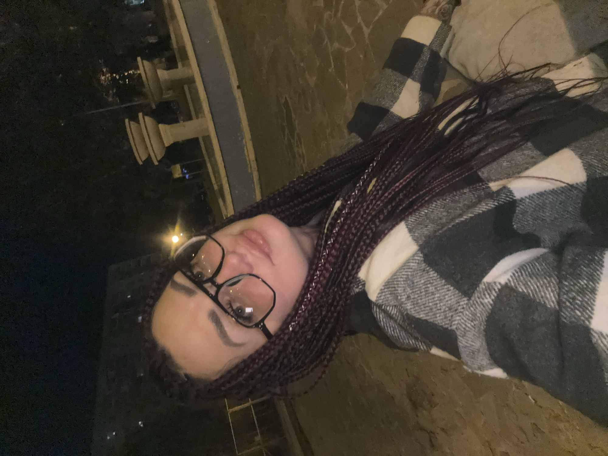 A woman with braided hair and glasses takes a selfie outdoors at night in Tolyatti, Russia. She is wearing a plaid jacket. In the background, there is a fountain and a building. The ground is paved with stones.