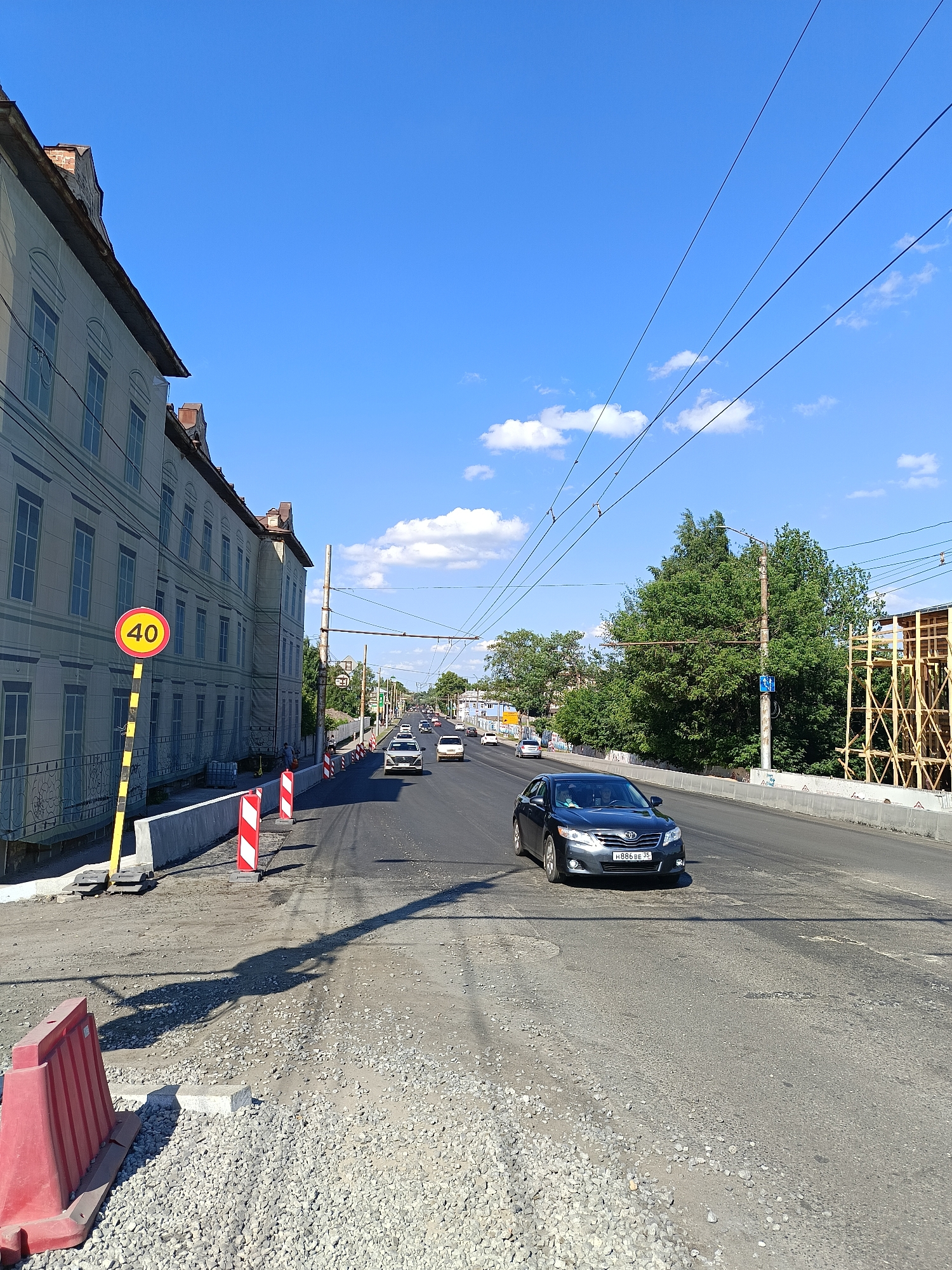 The image depicts a street scene in a city, likely undergoing some form of construction or repair. On the left, a large, multi-story building with a light-colored facade and arched windows lines the street. A yellow pole with a circular sign displaying "40" in red is positioned in front of the building. The street itself is paved, with visible signs of repair work, including loose gravel and what appears to be an unfinished road surface in the foreground. Several cars are present on the road, with one dark sedan in the foreground and others further down the street. Red and white striped traffic cones are placed along the side of the road, indicating a temporary hazard or work zone. Overhead, power lines crisscross the blue sky, which is dotted with scattered white clouds. Trees are visible on the right side of the road, and some scaffolding is also present. The overall impression is of an active urban environment with ongoing infrastructure work.