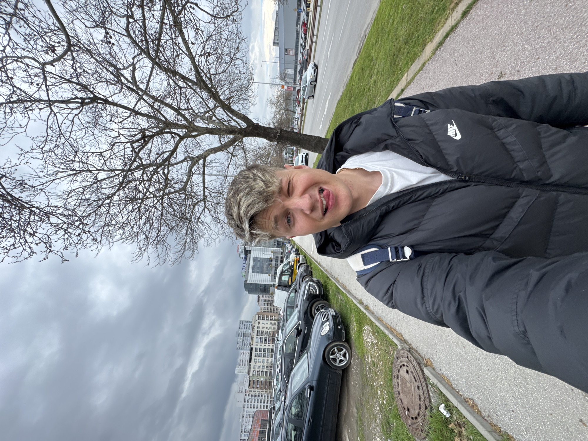 A young male is depicted in a selfie on a cloudy day in Plovdiv, Bulgaria. The subject has light-colored hair, blue eyes, and is wearing a black Nike puffer jacket over a white t-shirt with a visible gold-colored chain. He is winking with his right eye and sticking out his tongue, holding the camera in front of him. A blue and white backpack strap is visible over his right shoulder. He stands on a light-colored paved sidewalk, which features a circular metal manhole cover and a discarded white plastic cup in the lower left. To the subject's left, beyond a concrete curb, is a dirt parking lot occupied by numerous parked vehicles, including several dark sedans, SUVs, and a yellow and black recovery truck. In the background to the left, multi-story residential buildings are visible, alongside a modern glass-fronted office building bearing the "TELUS" logo and a red commercial sign. To the subject's right, a strip of green grass borders a multi-lane asphalt road with white lane markings, where several vehicles are in motion, including a silver hatchback. A large, leafless deciduous tree stands between the sidewalk and the road. Further in the background, across the road, a large grey commercial building is visible, featuring a prominent yellow McDonald's arch sign. The sky is overcast with uniform grey clouds. The location is identifiable as an urban area in Plovdiv, Bulgaria, specifically near the TELUS International office building and the Plovdiv Plaza shopping center.