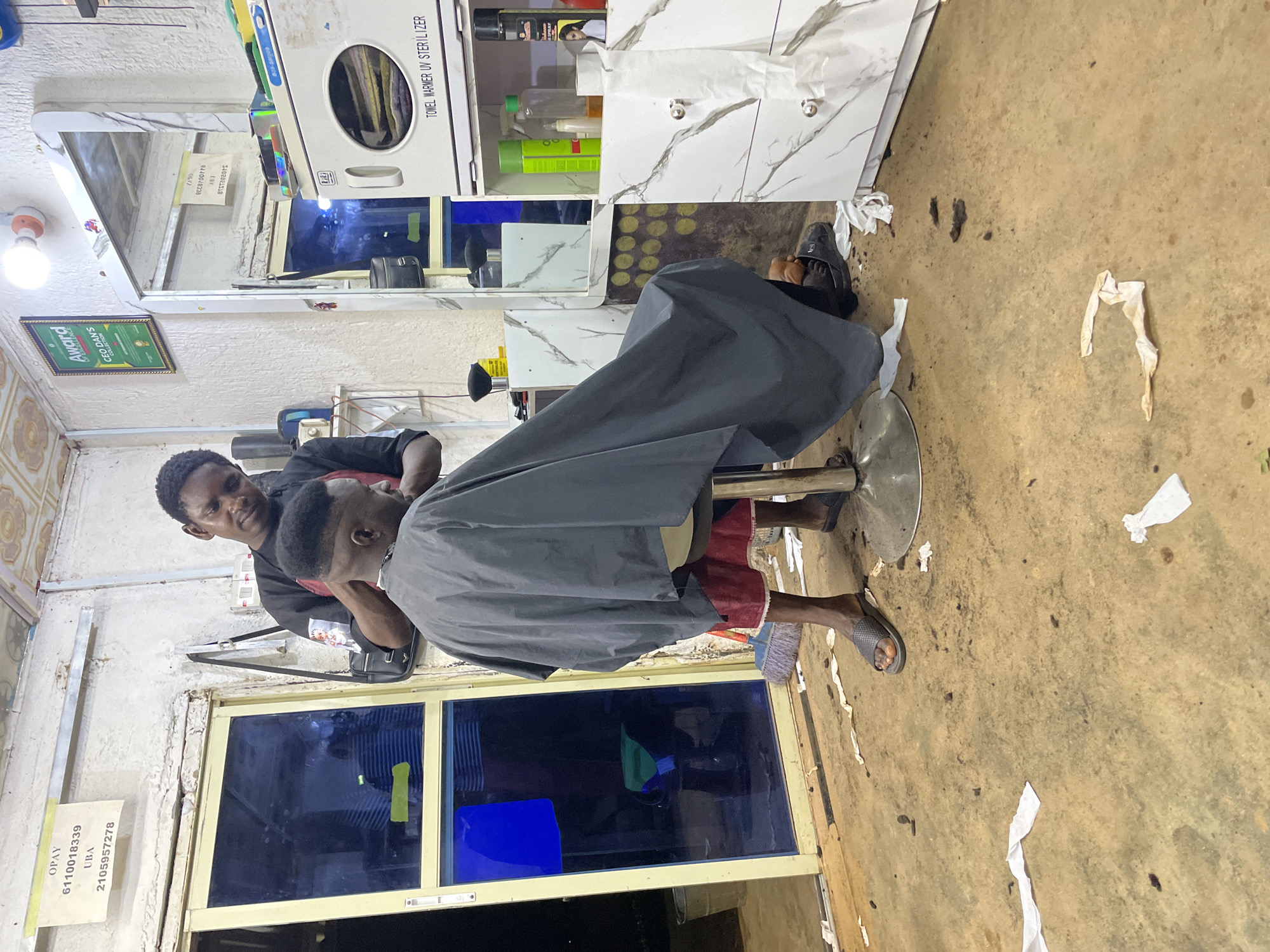 The image depicts an indoor scene at a barbershop in Durumi, Nigeria. A barber, wearing a black shirt with a red apron, is actively cutting the hair of a customer seated in a barber chair. The customer is draped in a grey cape, covering their body. The floor of the barbershop appears to be made of concrete and is littered with cut hair and paper scraps, suggesting a recent haircut.
To the left of the scene, a blue-tinted glass door is visible, with a yellow frame. Above the door, a sign with "OPAY" and account numbers is affixed to the wall. On the right side of the image, a white cabinet with marble-patterned doors and silver knobs is present. On top of this cabinet, various items are displayed, including a green spray bottle, a clear bottle with liquid, a roll of white paper towels, and a towel warmer/UV sterilizer unit. A mirror is mounted above the cabinet. Further on the wall, a framed award labeled "Award CEO DAN'S COLLECTION" is displayed.
The lighting in the barbershop appears to be from overhead bulbs. The overall environment suggests a functional, albeit somewhat cluttered, barbershop.