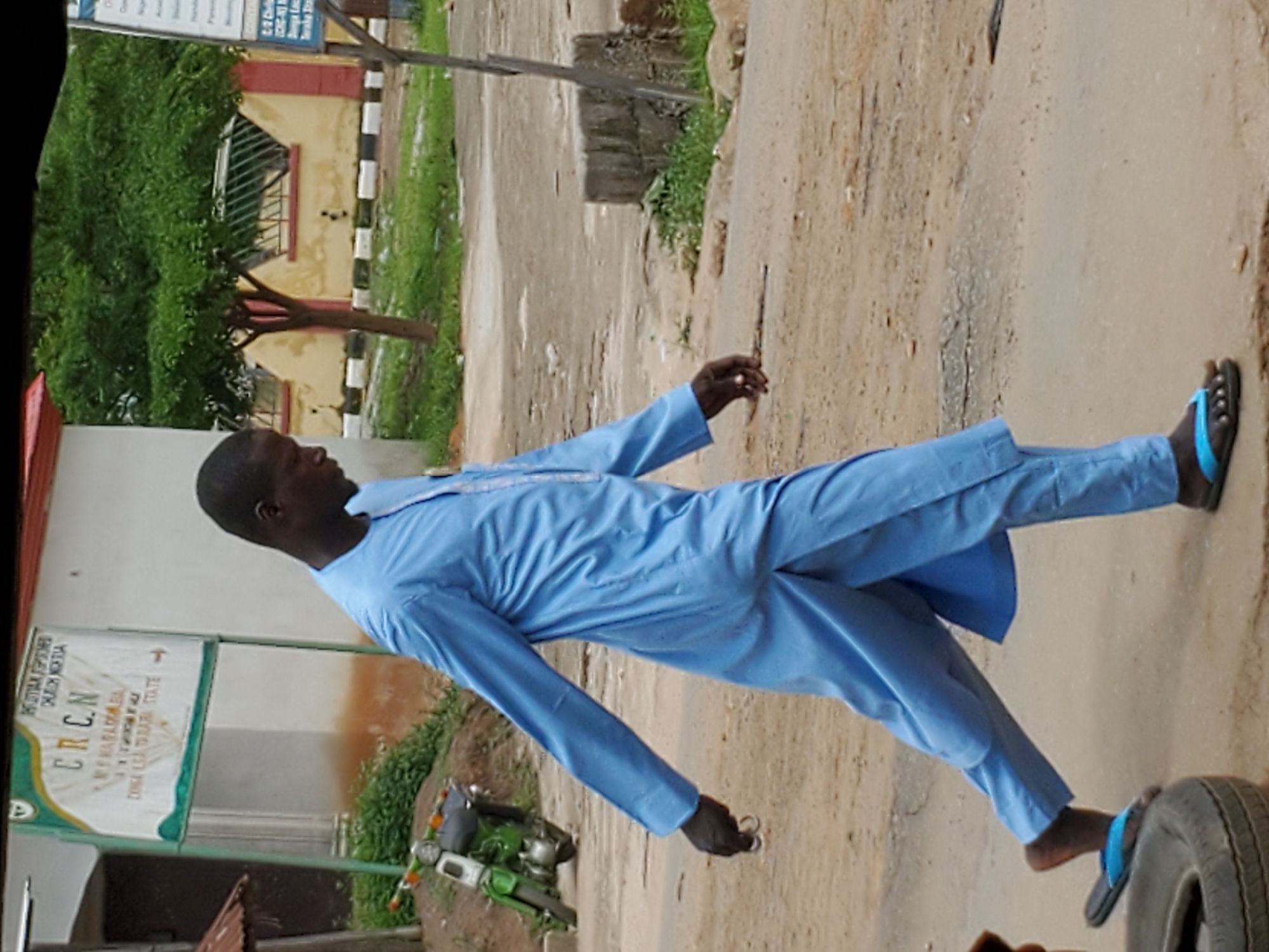 This outdoor, daytime full-shot captures a man standing on an unpaved road in Mararaba, Nigeria. The man, of African descent, is dressed in a light blue traditional tunic and matching trousers, complemented by black flip-flops with blue straps. He holds a small, metallic, circular object in his right hand, possibly keys, and is looking towards his left.
The setting appears to be a semi-urban or rural pathway. The ground is a mix of dusty, sandy earth and some concrete, with patches of green grass and foliage visible. A green motorcycle or scooter is partially visible on the left side of the frame. A prominent white sign with green borders stands near the ground on the left. The text on the sign reads: "CHRISTIAN REFORMED CHURCH OF NIGERIA", "CRCN", "CRCN HAFADEMA", and "DIMA LEA TARABA STATE", firmly placing the location within Taraba State, Nigeria.
In the background, a light yellow building with red trim around a window and a distinctive black and white striped barrier is visible. Lush green trees and shrubs are scattered throughout the scene. The natural lighting suggests it is daytime, likely late morning or afternoon, with no extreme shadows. The man's posture suggests he is standing or walking casually, with a neutral expression. A black tire is partially visible in the bottom right corner of the image.