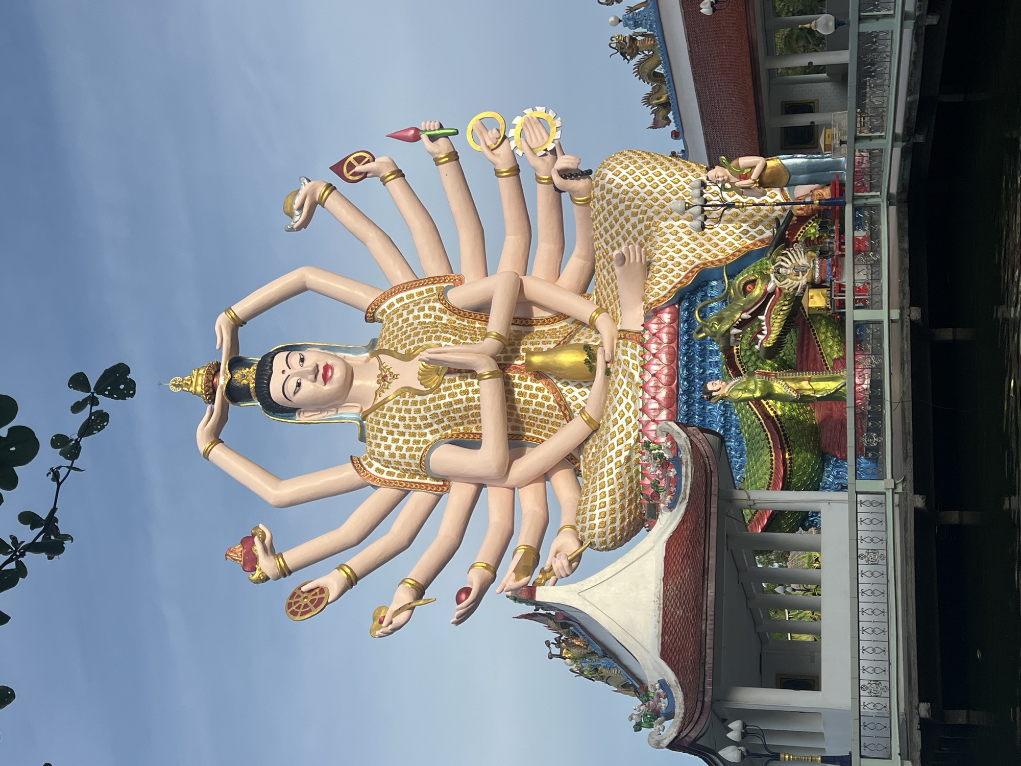 The image shows a large, multi-armed statue of a deity, likely Guanyin, in Baan Plai Laem, Thailand. The statue is seated and adorned in gold-patterned robes, with numerous hands holding various symbolic objects. Below the deity, a colorful dragon sculpture is prominently featured, adding to the vibrant and elaborate artistic style. The setting appears to be a temple or cultural complex, with traditional Thai architectural elements visible in the surrounding structures, including red-tiled roofs and ornate detailing. The scene is captured outdoors under a clear blue sky, with some tree branches visible in the upper portion of the frame.