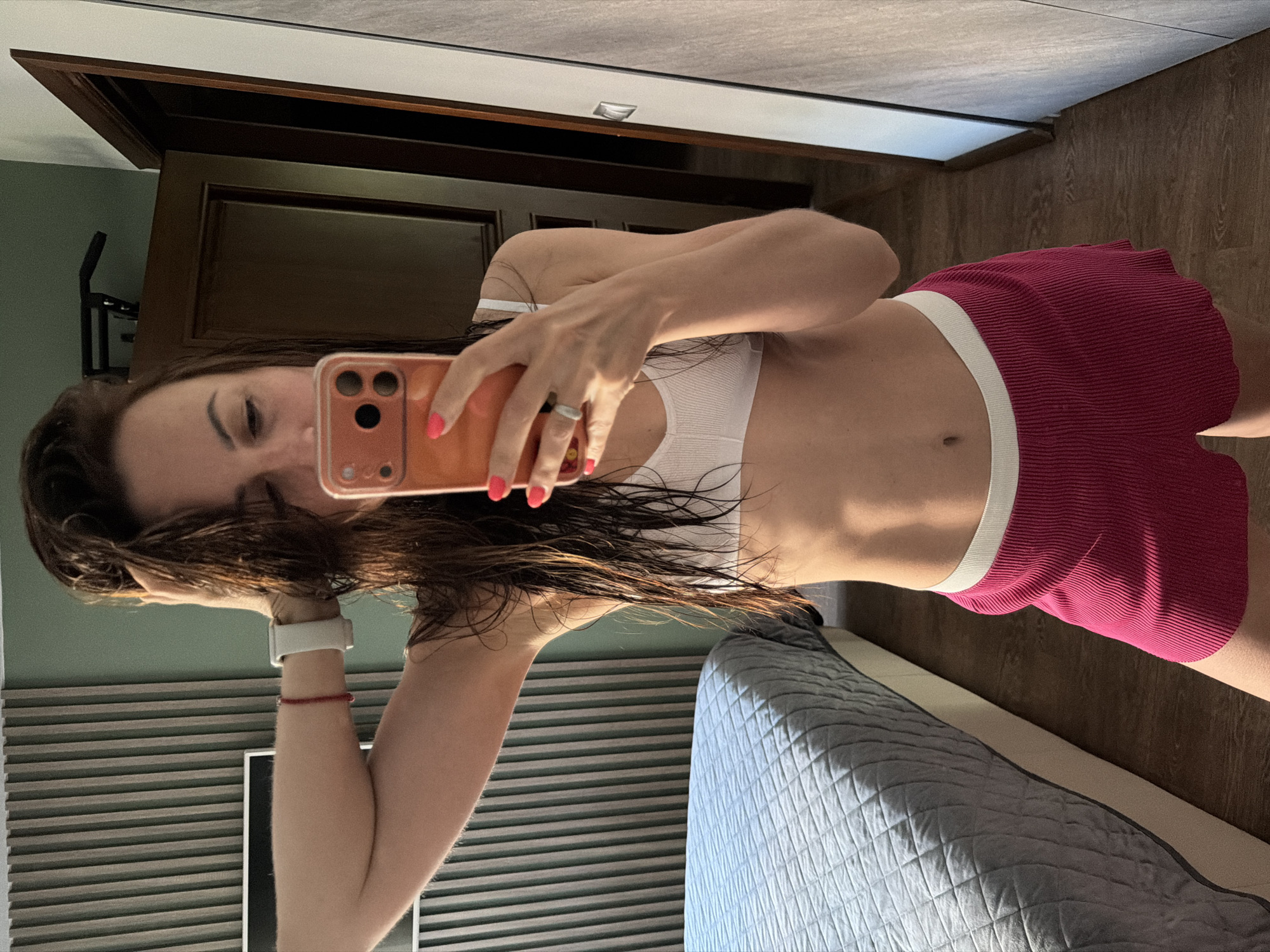 A woman is taking a mirror selfie in a bedroom, holding her phone with her right hand. She is wearing a white sports bra and pink shorts. Her hair is wet and she is touching it with her left hand. The room has a bed with a grey quilted comforter, a wooden floor, and a wall with vertical wooden slats. A door and a closet are visible in the background. The scene appears to be indoors, likely in a residential setting.