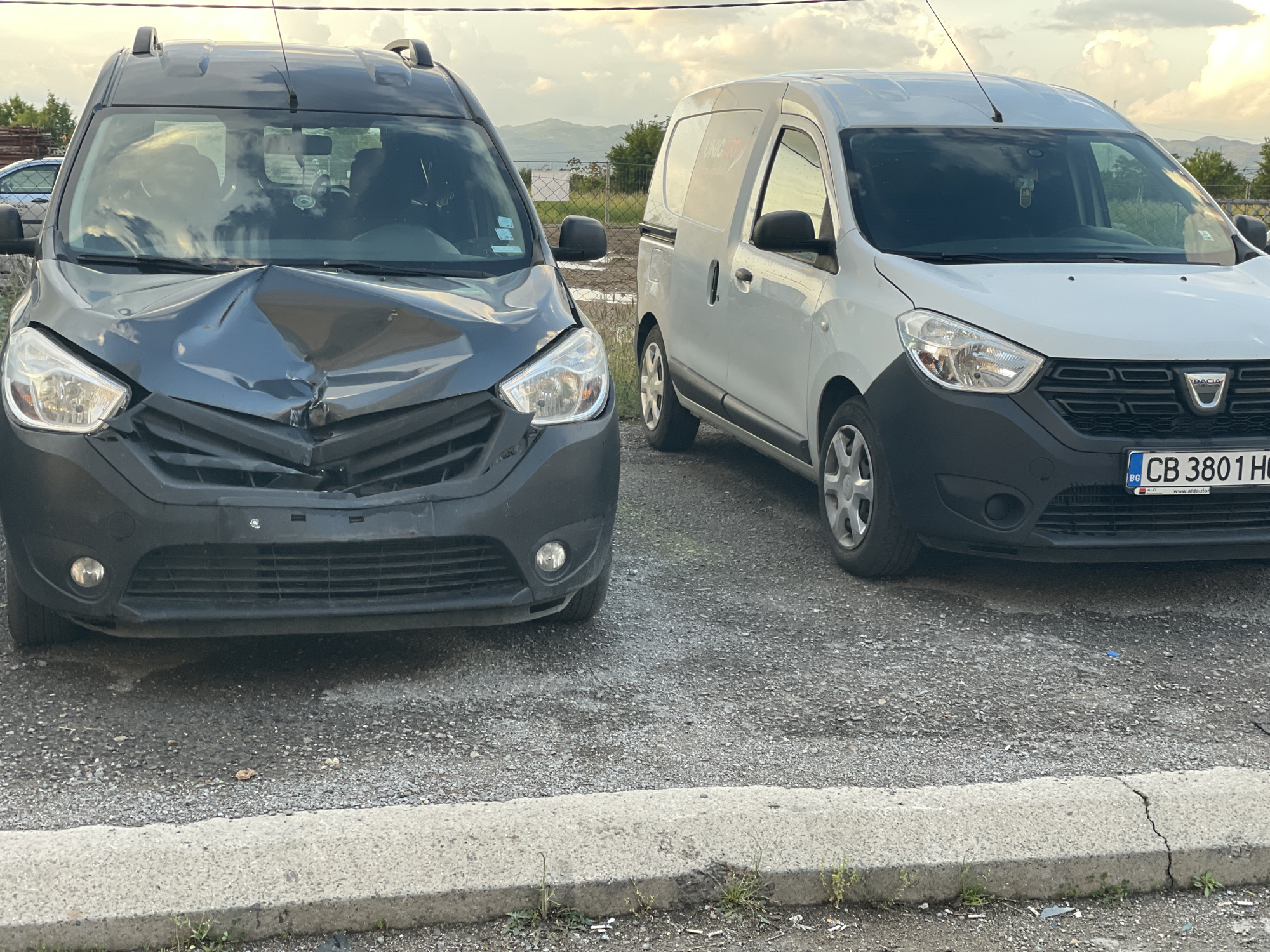 The image shows two vans parked in what appears to be a parking lot. The van on the left is dark grey and has significant front-end damage. The van on the right is white and appears undamaged. The license plate on the white van is visible and indicates it is from Bulgaria. The background includes buildings, vegetation, and a cloudy sky. The setting is an outdoor area, possibly a roadside or commercial area.