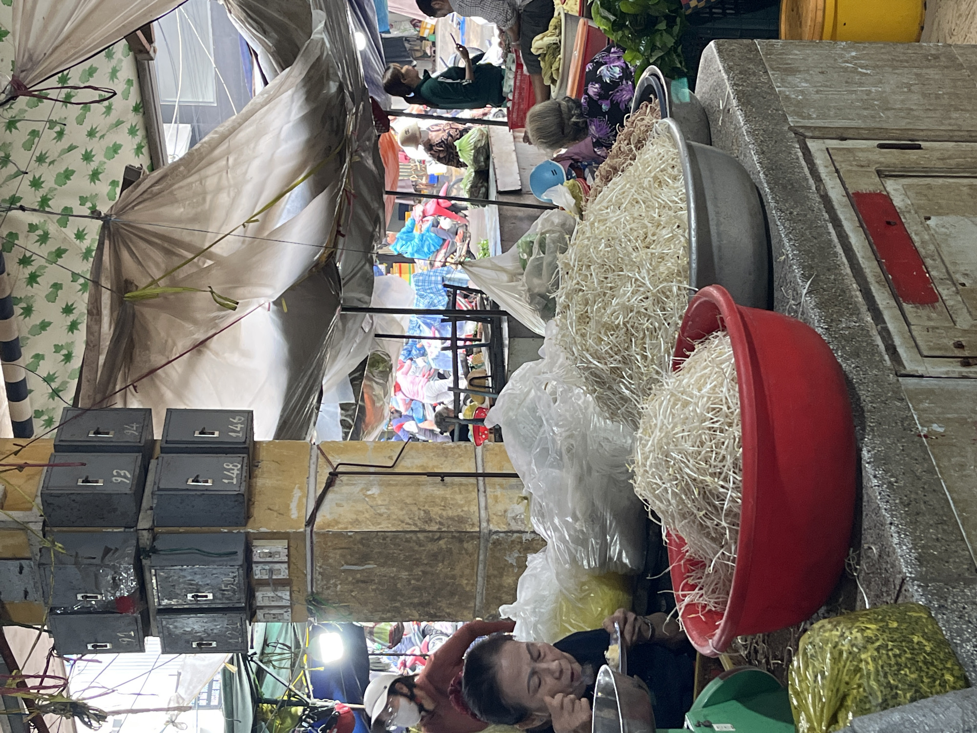This media file depicts a busy, covered market scene in Bac Nha Trang Ward, Vietnam. The image is oriented vertically (rotated 90 degrees clockwise from its natural upright position) but described here as if viewed upright.
In the immediate foreground, large quantities of bean sprouts are piled in a grey basin and a bright red bowl, indicating a fresh produce stall. A woman with dark hair is visible, looking downwards and eating from a bowl with a spoon. A green weighing scale and a bag of green leafy produce are also in this area.
The mid-ground reveals a concrete counter or stall surface. Above it, several grey metal boxes are stacked, secured to a beam or support structure. Some of these boxes have visible numbers: "94", "148", "93", "148", "91", and "421". The market space is covered by an assortment of tarps, awnings, and plastic sheets, providing shade or shelter. A patterned fabric with green leaf motifs is visible on the left.
Further into the background, other people populate the market. One woman in a green top is seen resting or using a phone. More individuals are scattered throughout the stalls, suggesting various activities like selling, buying, or simply waiting. The background shows a dense arrangement of market goods, possibly clothing or other produce, under diverse covers. A yellow bucket is discernible in the upper right section.
The lighting appears to be a mix of natural diffused light coming through the coverings and artificial light from a fixture visible on the lower left. The overall scene conveys a lively, traditional market atmosphere.