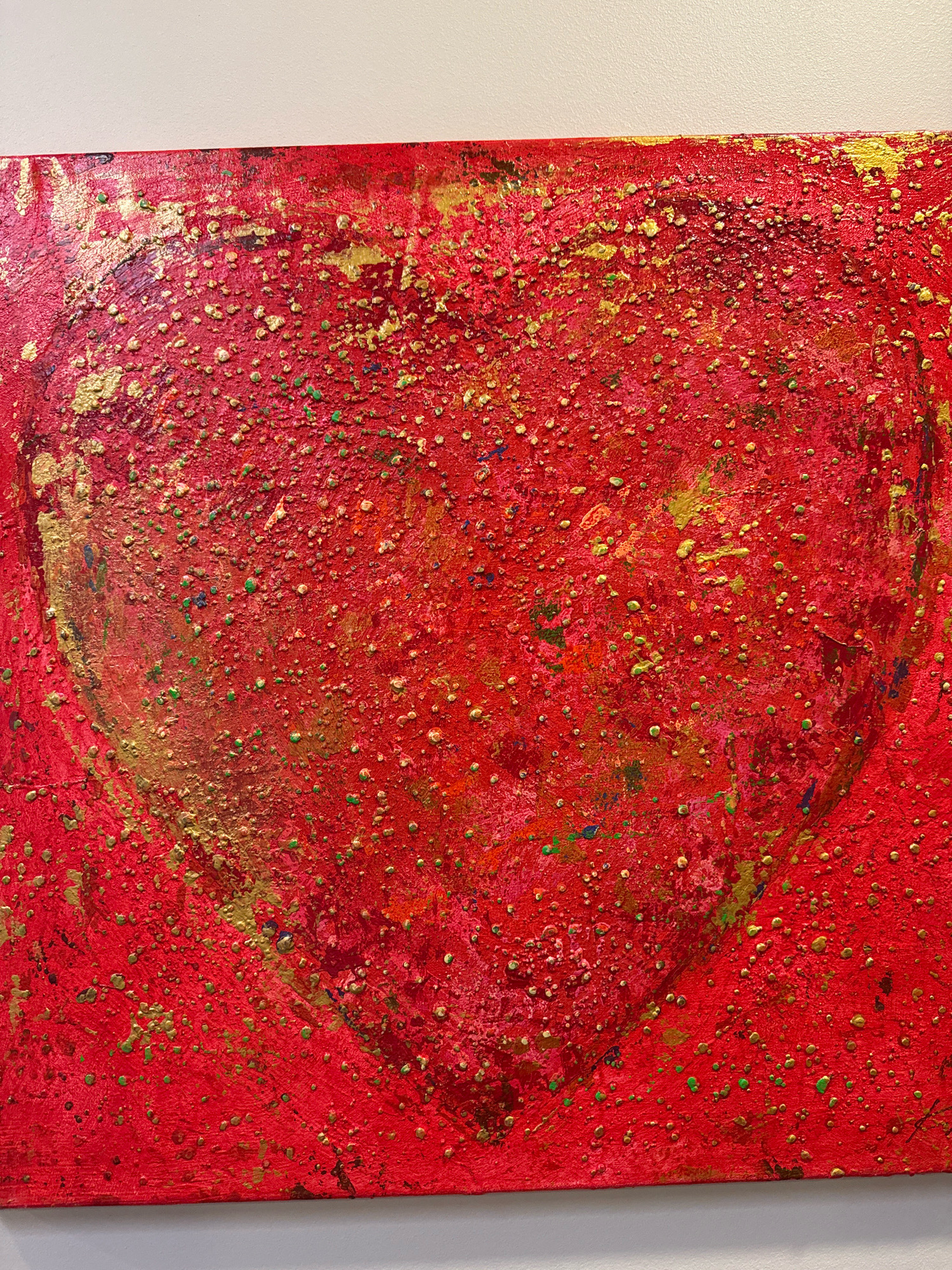 The image displays a close-up, eye-level shot of a textured red abstract painting featuring a large, indistinct heart shape. The artwork is rendered on a canvas and is predominantly red, with various specks and splatters of gold, green, and other colors scattered across its surface. The texture appears raised and uneven, suggesting a thick application of paint or the use of mixed media. The painting is mounted on a wall against a plain, off-white background. There are no people, animals, or recognizable objects visible within the frame, besides the painting itself. The location in Tallinn, Estonia, is not discernible from the visual information provided in the image.
