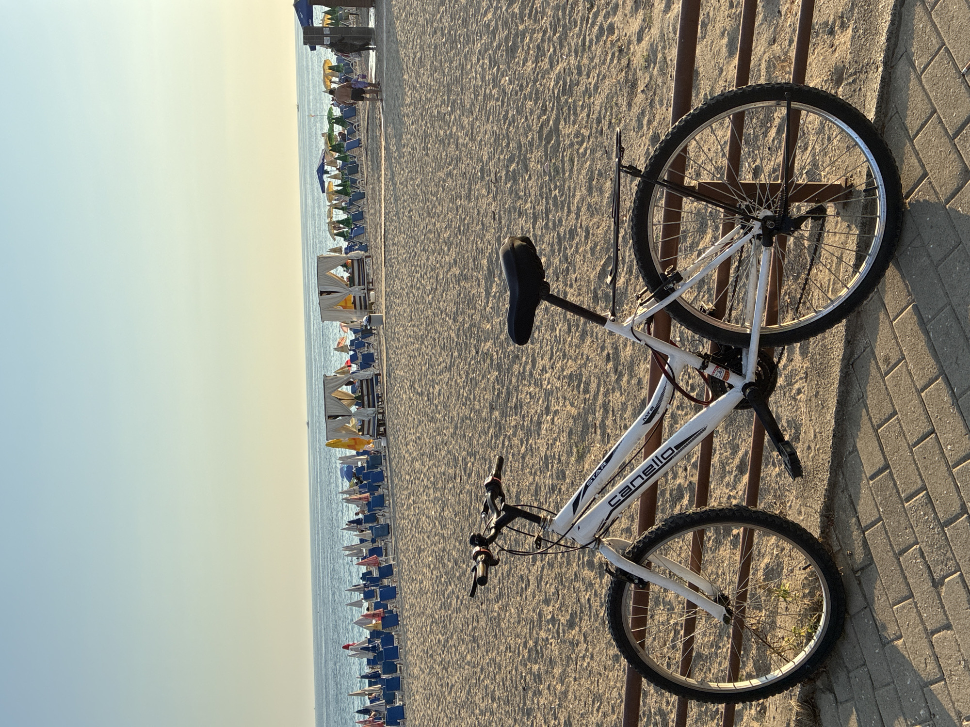 A white bicycle is parked on a brick walkway in front of a sandy beach lined with rows of blue lounge chairs and colorful umbrellas. The sea is visible in the background under a clear, pale blue sky. A few people are walking on the beach in the distance. The scene appears to be a public beach in Alanya, Turkey, likely during daytime hours given the lighting.