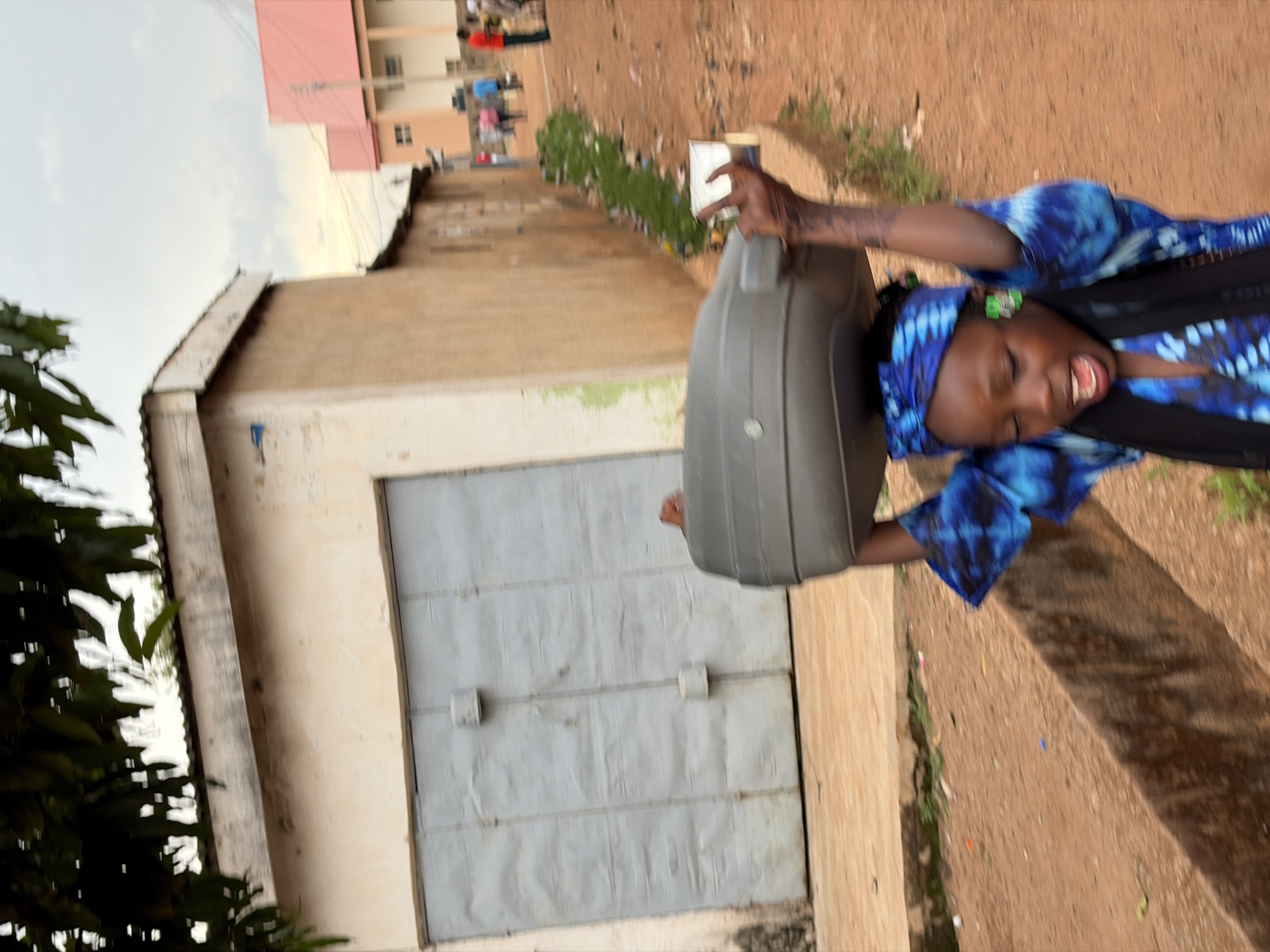 A young woman wearing a blue tie-dye patterned top and headscarf is carrying a large gray container outside a building with gray metal doors. The scene is likely in Jos, Nigeria, as inferred from visual cues suggesting an urban environment in that region. Additional buildings and people are visible in the background, situated higher than the primary scene. The ground is dirt.