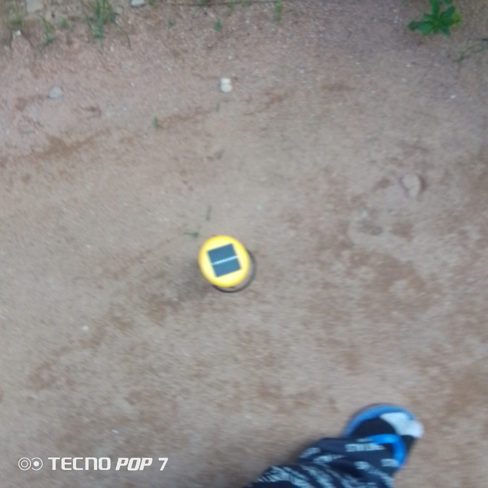 The image shows a close-up, top-down view of a solar-powered garden light on a dirt path. The light is round and yellow with a dark blue square solar panel on top. The path is sandy and there are a few blades of green grass scattered around. In the bottom right corner of the image, a person's foot wearing a blue and black flip-flop is visible. The image is slightly blurry, suggesting it might have been taken with a phone camera. The text "TECNO POP 7" is visible in the bottom left corner, indicating the phone used to take the picture. The scene appears to be outdoors, likely during the day given the solar light. The setting is informal and appears to be a walkway or garden area in Jalingo, Nigeria. There is no specific activity or event taking place other than the placement of the solar light on the path.