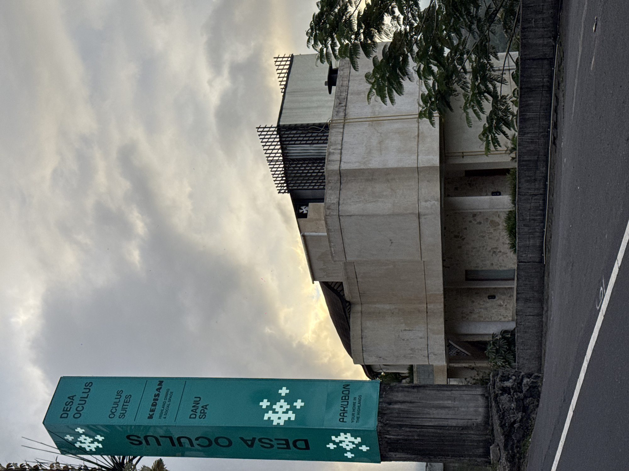 A teal-colored signpost for "Desa Oculus" stands tall on the left side of the frame, displaying various services like "Oculus Suites," "Kedisan Highland Dining & Social Space," and "Danu Spa." The signpost also features a white emblem of interconnected plus signs. Behind the signpost, a concrete building with stone accents is visible, leading to an upper structure with a distinctive latticework facade. The sky above is overcast with diffused sunlight, suggesting either early morning or late afternoon. The scene appears to be set in Kintamani, Indonesia, possibly a resort or complex. The foreground shows a paved road with a white line marking.
