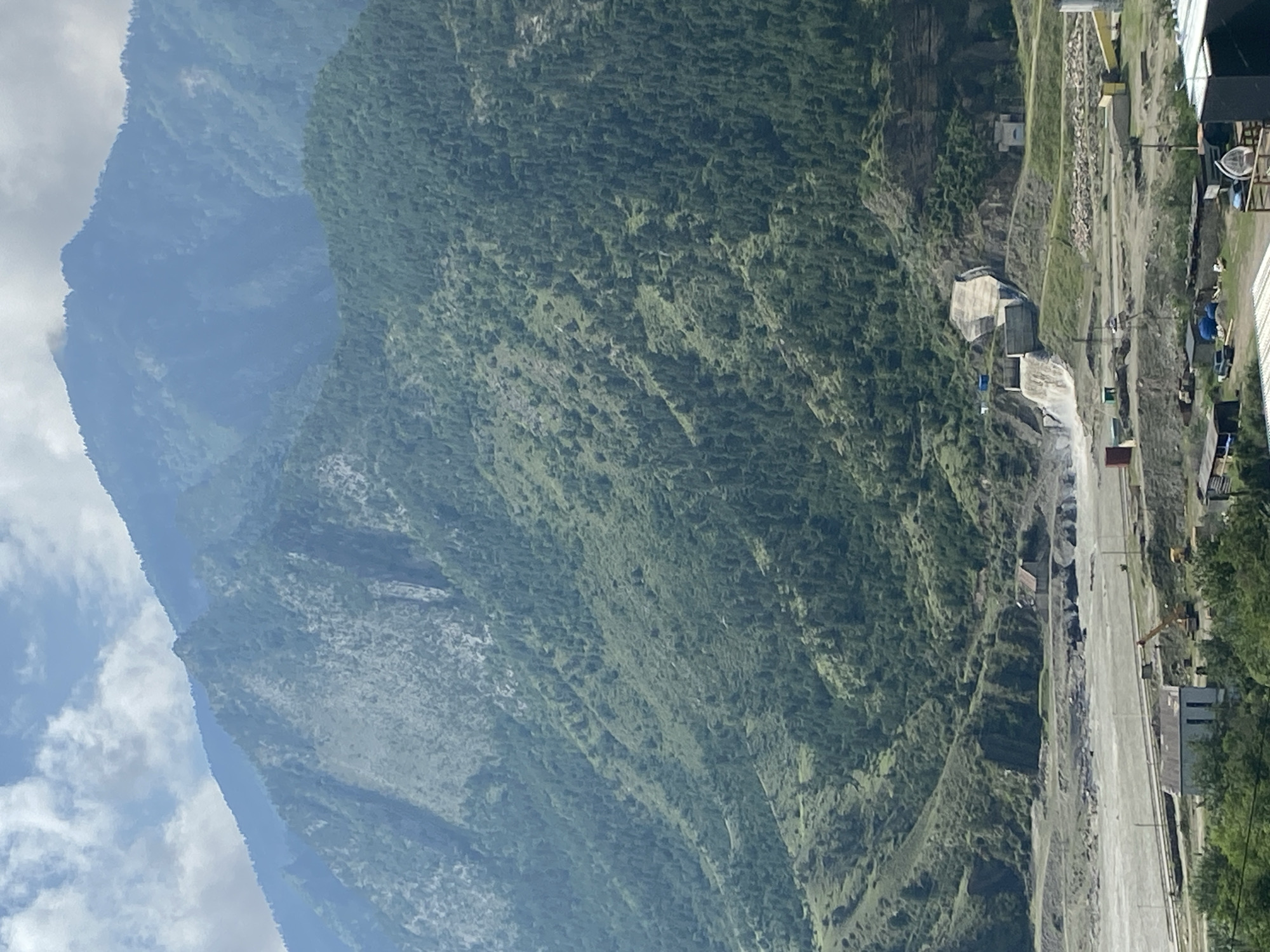 The image depicts a mountain landscape with a significant man-made structure, likely a hydroelectric dam, built into a steep hillside. Water cascades down the concrete spillway of the dam, flowing into a river or reservoir below. The surrounding mountains are covered in dense green vegetation, with a mix of trees and scrub. The sky above is partly cloudy. In the foreground, there is evidence of construction or industrial activity, including buildings, construction equipment such as a crane and excavators, and various storage containers. The overall scene suggests a remote, mountainous region where infrastructure has been developed to harness natural resources. The location is identified as Chalda, Russia, a village known for its proximity to natural landscapes and potential for hydropower development.