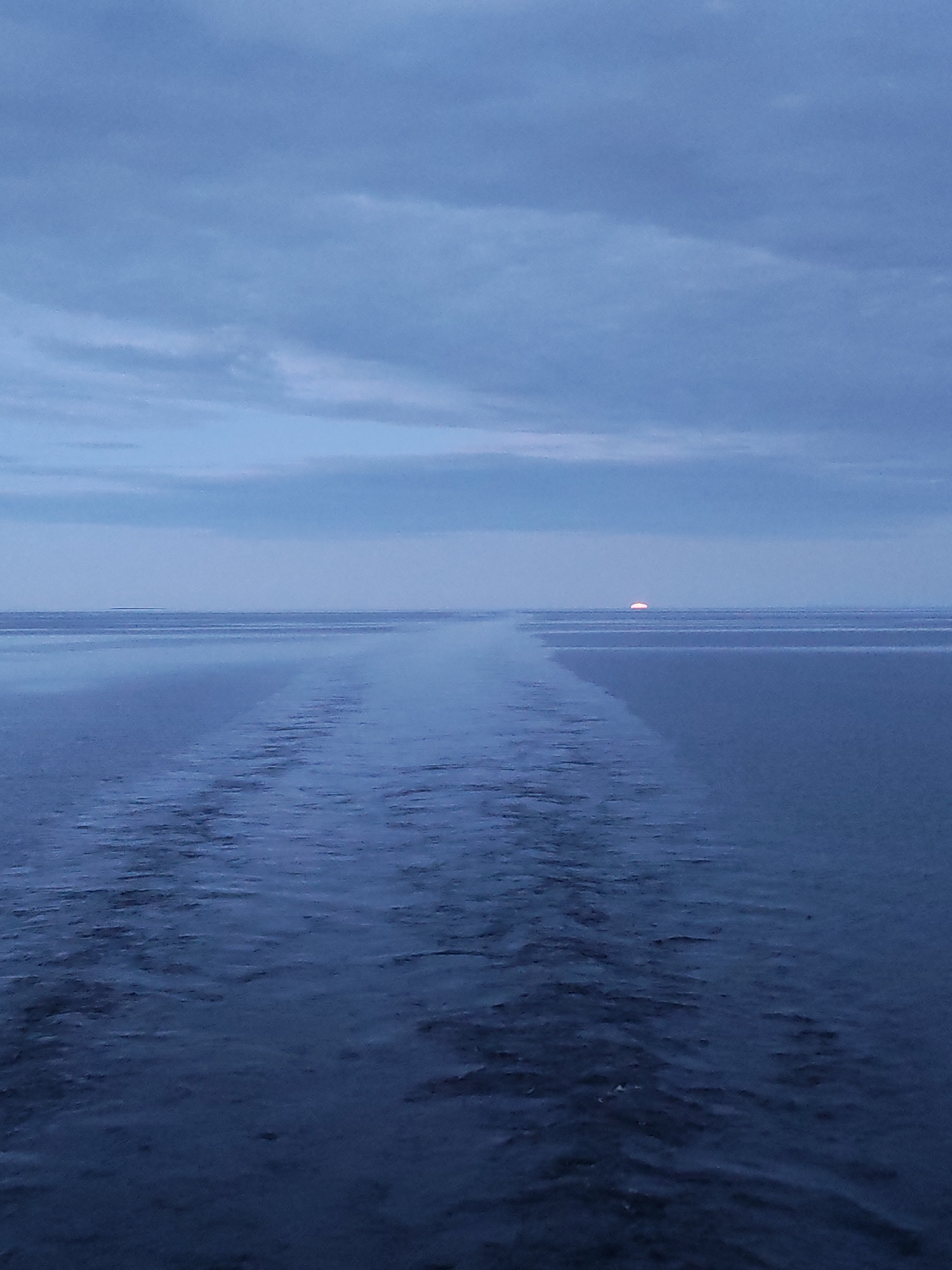 The image presents a wide, serene vista of open water under a cloudy sky, likely captured during dawn or dusk. The dominant colors are various shades of cool blue and grey, creating a muted and atmospheric scene.
In the foreground and midground, the surface of the water shows a distinct wake or trail, suggesting the photograph was taken from a moving vessel. This disturbance in the water extends in a relatively straight line towards the distant horizon, composed of ripples and smoother patches.
The horizon line is very clear and stretches horizontally across the image, separating the water from the sky. Just above the center of this horizon, a small, luminous semicircle of orange-red light is visible, indicating the sun or moon partially rising or setting.
The sky is filled with broad, soft clouds that appear layered and diffuse, ranging from lighter blue-grey near the horizon to darker tones higher up. The overall weather appears calm, with no signs of turbulence or heavy precipitation.
There are no people or discernible man-made objects apart from the implied vessel creating the wake. No visible text is present in the image. The scene conveys a sense of vastness and tranquility, characteristic of a remote maritime or large lake environment, with the given context of "Unknown, Russia."