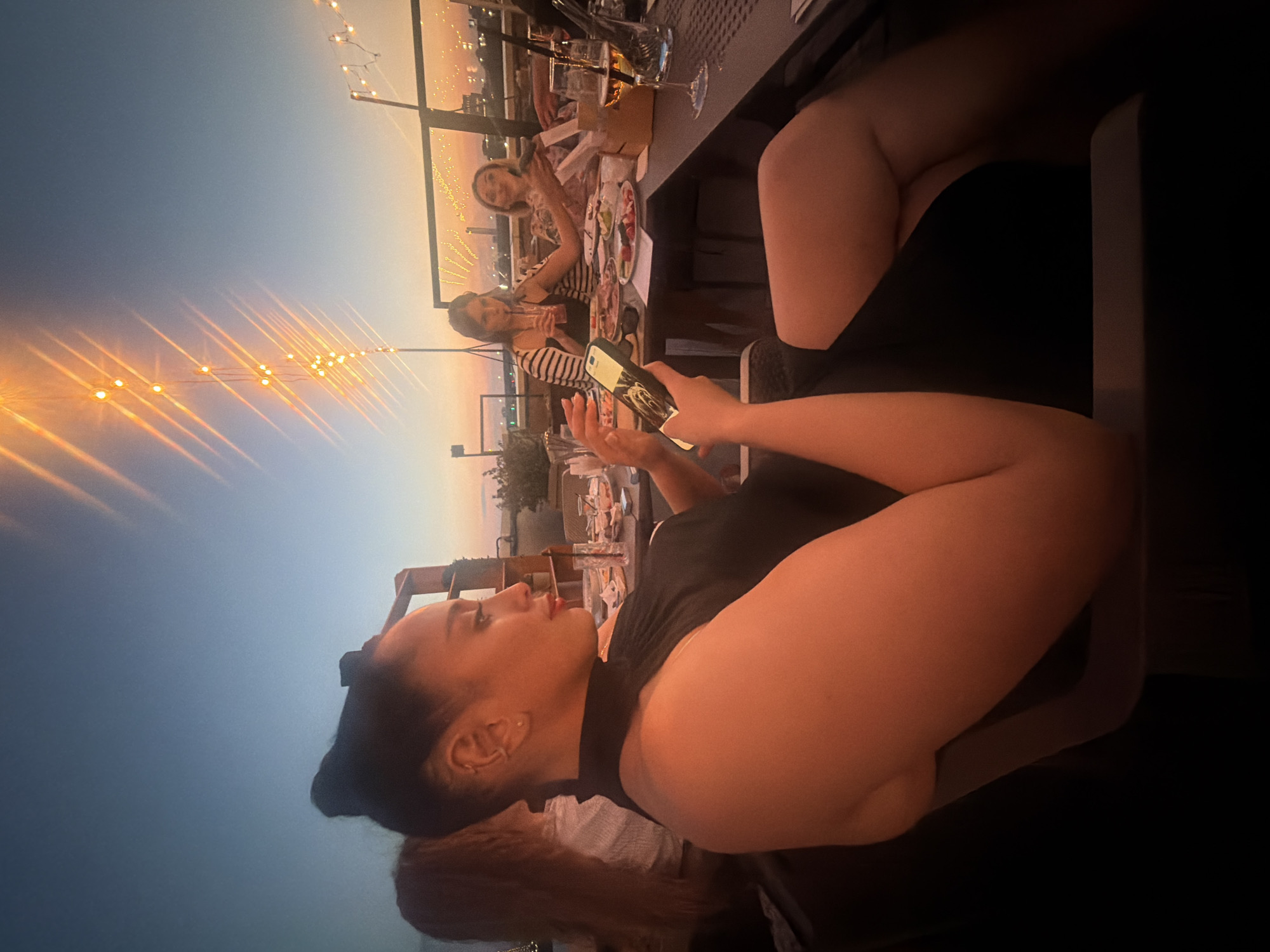 This media file depicts a group of people enjoying a meal at an outdoor restaurant during twilight. The subjects are primarily women, seated around a table laden with food and drinks. One woman in the foreground is looking away from the camera, her arm resting on the table. Another woman, seated further back, is holding a phone and looking at it. The setting appears to be a rooftop or elevated patio, suggested by the view of distant lights and the presence of string lights overhead. The time of day is evident from the gradient of the sky, transitioning from a pale blue to a warmer, orange hue near the horizon, indicative of sunset or dusk. The mood seems relaxed and social, with people engaged in conversation and enjoying their meal. No visible text is present in the image. The context suggests this scene may be taking place in Novosibirsk, Russia.