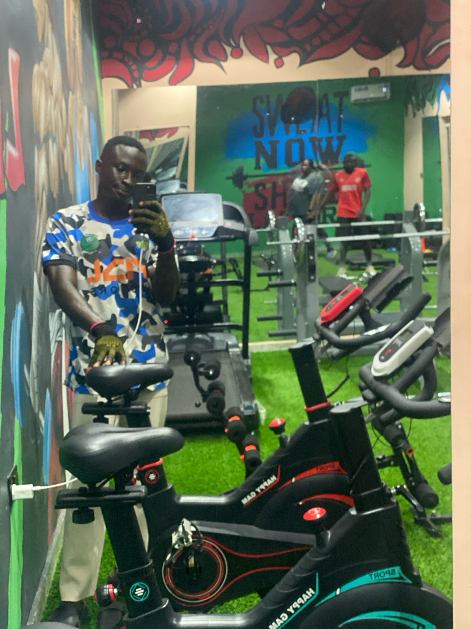 A dark-skinned man, wearing a blue and camouflage t-shirt and light-colored pants, is taking a selfie in a gym. He is holding a black smartphone in his right hand and wearing green fingerless gloves and pink wristbands. He is standing in front of a stationary bicycle. The gym has artificial turf flooring and mirrors on the walls, one of which reflects the man and the gym equipment. In the background, visible in the mirror, are other gym-goers, weightlifting equipment, and motivational graffiti on the wall that reads "SWEAT NOW SHINE LATER". There are also treadmills and other exercise bikes. The environment is brightly lit.