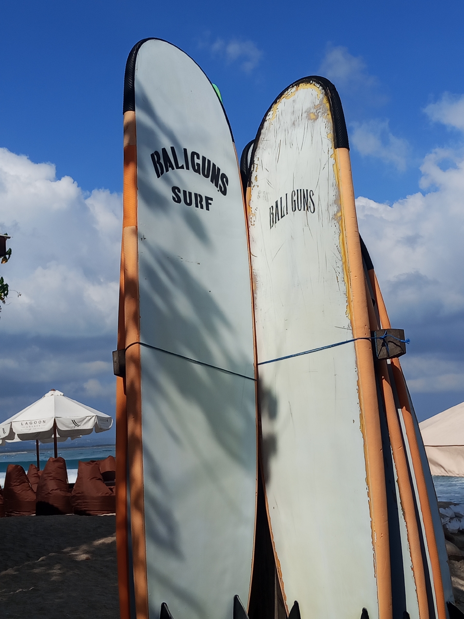 Two surfboards, weathered and bearing the branding "BALIGUNS SURF" and "BALI GUNS," stand upright on a sandy beach under a bright blue sky with scattered clouds. The surfboards have orange edges and are leaned against each other. A blue rope is tied around the right surfboard and a dark block. In the background, a white umbrella with the word "LAGOOON" is visible, along with several brown beanbag chairs and the ocean. The scene depicts a typical beach setup in Kuta, Indonesia, likely a surf school or rental location. The sand appears dark, suggesting it could be volcanic sand common in some parts of Bali.
