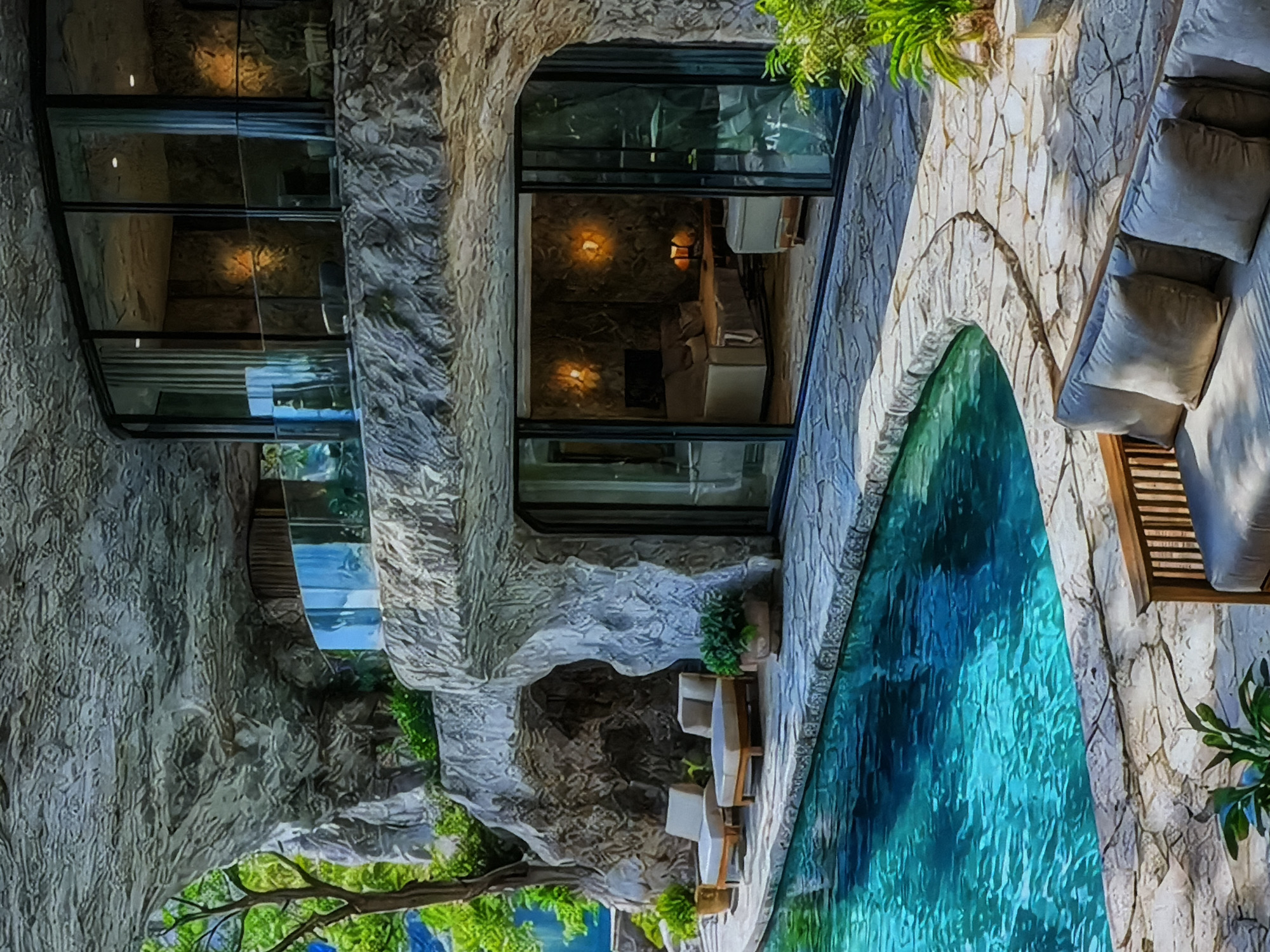 The image depicts a luxurious architectural complex, likely a villa or resort, characterized by its distinctive design that integrates with a rocky or cave-like structure. The main building on the left features a multi-story facade with expansive, dark-framed glass windows and sliding doors, revealing glimpses of the interior. The exterior surfaces are rough and textured, mimicking natural stone or rock formations.
Adjacent to the building, a large, irregularly shaped swimming pool with vivid blue water dominates the right side of the frame. Surrounding the pool is a patio paved with light-colored stone, furnished with outdoor seating. This includes a long sofa adorned with light-colored cushions and pillows, and further down, a pair of lounge chairs with a small table. Green foliage and potted plants are interspersed throughout the scene, enhancing the naturalistic aesthetic.
Through the large windows, interior spaces are visible, illuminated by warm, inviting lights. These include what appears to be a bedroom with a bed and a living area with furniture, suggesting comfort and modern amenities within the unique rock-hewn structure.
The scene conveys a sense of tranquility and luxury, likely set during the evening or night, given the prominence of artificial interior lighting and the absence of direct sunlight. No people are visible, and there is no discernible text in the image. The location is Sofiivska Borshchahivka, Ukraine.