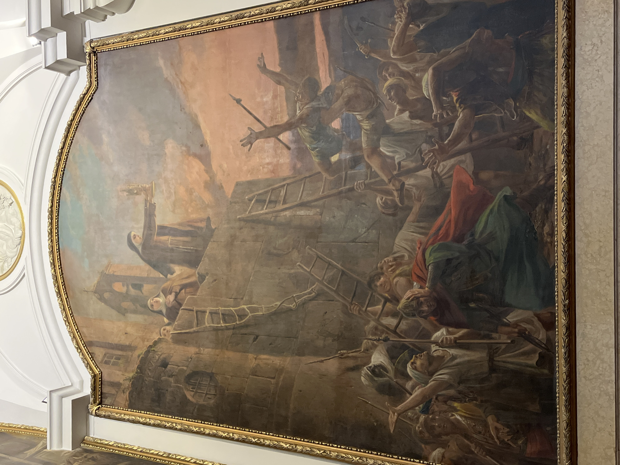 The media file displays a large, dramatic oil painting depicting a historical siege scene, encased in an ornate gold frame and mounted on a white wall, likely in a building in Buenos Aires, Argentina.
The painting's composition is sharply divided between a defending party positioned atop a fortified stone wall or city, and an attacking force attempting to scale it from below. On the left side, several figures in religious habits, likely nuns, are seen on the ramparts. One prominent nun in the upper-left corner holds a golden monstrance or similar religious relic aloft, seemingly invoking divine protection against the encroaching attackers. Other religious figures appear watchful or concerned, looking down at the melee.
Below and to the right, a tumultuous mass of men, comprising the attacking force, struggle to ascend the wall using multiple wooden ladders. They are depicted with dynamic, aggressive poses, some brandishing spears, swords, and clubs. Several figures are mid-climb, while others appear to be falling or pushing forward. Their attire suggests a historical period.
The sky in the background features warm, fiery orange and red tones blending into cooler blues and greys, suggesting either dawn or dusk, and adding to the dramatic tension of the scene. The overall activity portrays a desperate struggle for the fortified location. The emotions conveyed range from the attackers' aggression and determination to the defenders' faith and perhaps fear. No discernible text is visible within the painting itself.