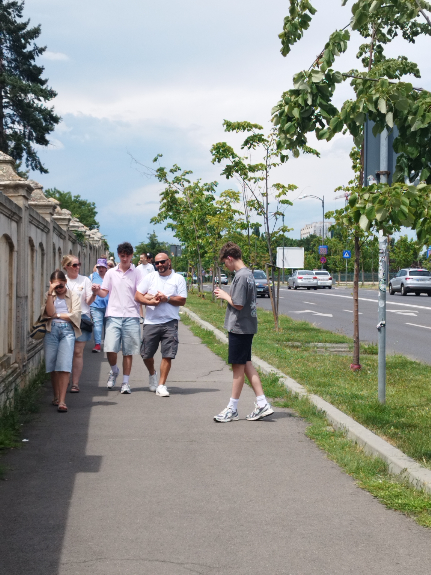 A group of people walk along a sidewalk next to a street in Bucharest, Romania. The group includes at least six individuals, walking in loose formation. A man in a light pink polo shirt and shorts walks with a young woman wearing a tan jacket over a white top and denim shorts, and a girl in a similar outfit. A man in a white t-shirt and grey shorts walks next to the man in the pink shirt, holding hands with a younger man in a grey t-shirt and black shorts who is looking at his phone. Other individuals are visible further back in the group. They are walking alongside a decorative stone wall on their left and a grassy area with trees and a street on their right. Cars are visible on the street. The sky is partly cloudy.