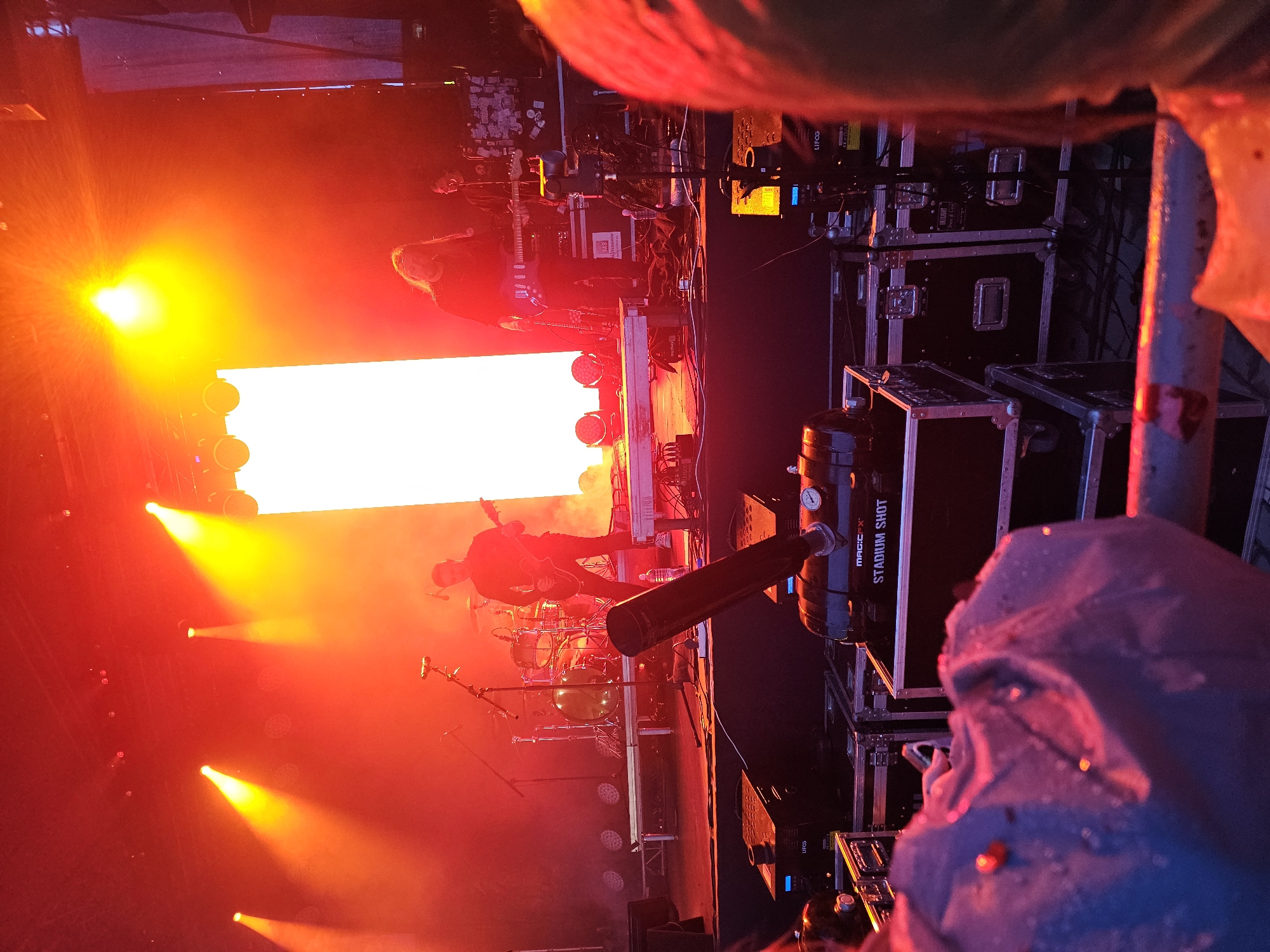 The image depicts an outdoor music concert in Kazan, Russia, characterized by intense red and orange stage lighting. In the foreground, there are large black equipment cases, one of which is labeled "STADIUM SHOT." A person in a blue raincoat is partially visible in the lower left corner. The stage features a drummer with a drum kit, a guitarist playing an electric guitar, and another person playing a keyboard or synthesizer. A bright, vertical white screen is positioned behind the performers. The overall atmosphere is dynamic and energetic, with smoke or haze contributing to the dramatic lighting effects.