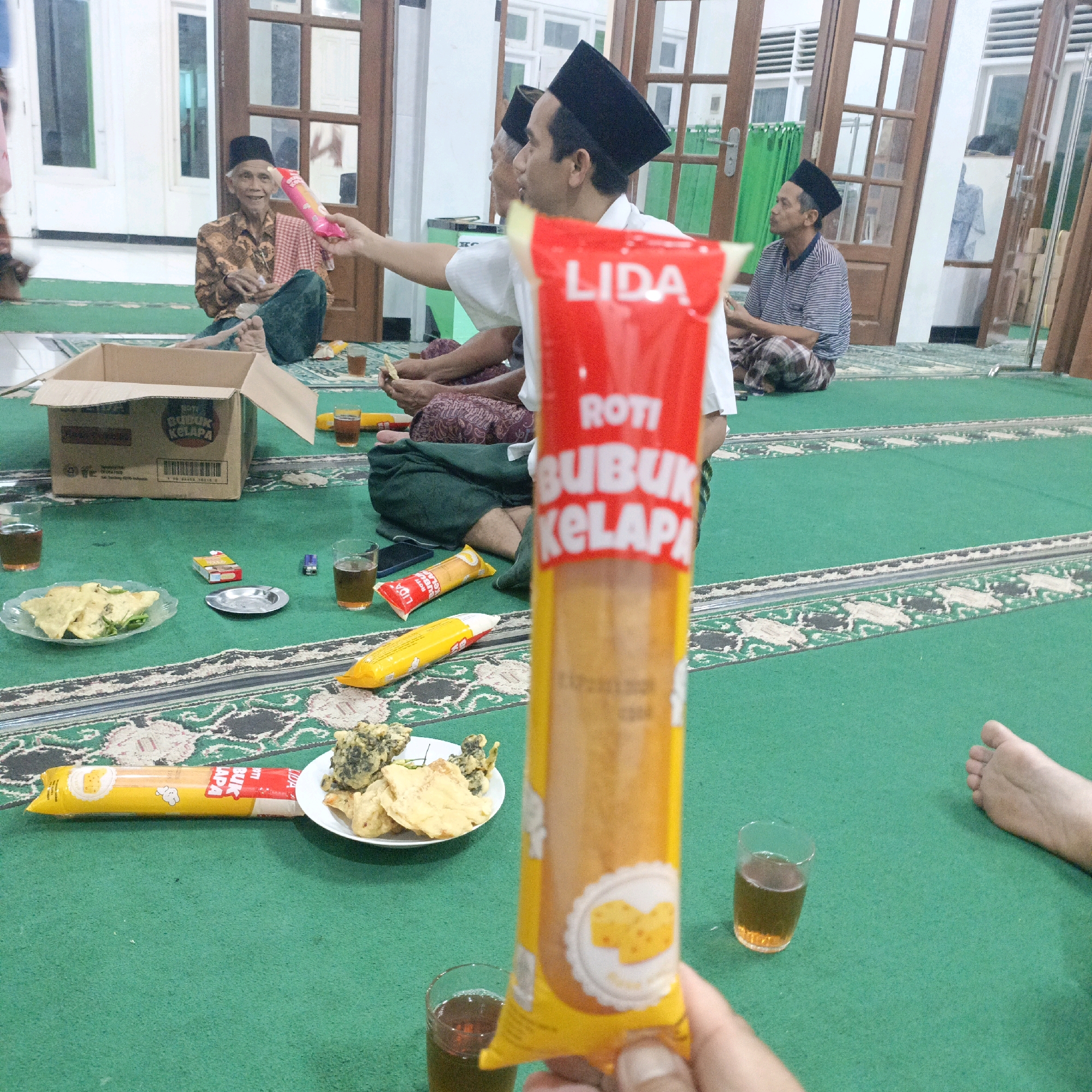 In Salatiga, Indonesia, a gathering is taking place on a green carpeted floor, likely inside a building with wooden doors and windows. The scene is informal, with several men seated on the floor. In the foreground, a person's hand is holding up a bright red and yellow package with Indonesian text that reads "ROTI BUBUK KELAPA," which translates to "COCONUT POWDER BREAD." This package is prominently displayed, partially obscuring the view of the other people and objects in the frame.
To the left of the foreground package, there is a plate of food, possibly fried items like tempeh or crackers, alongside a glass of dark liquid, likely tea. A cigarette pack and an ashtray are also on the carpet. Further into the background, an open cardboard box with text is visible, and another person is holding a similar food item. Several men are seated, some wearing traditional Islamic caps called peci. One elderly man in the background is smiling and looking towards the foreground. Another man is wearing a striped shirt. The environment suggests a communal gathering, possibly during a religious event or a casual get-together. The lighting appears to be natural, coming from the windows.