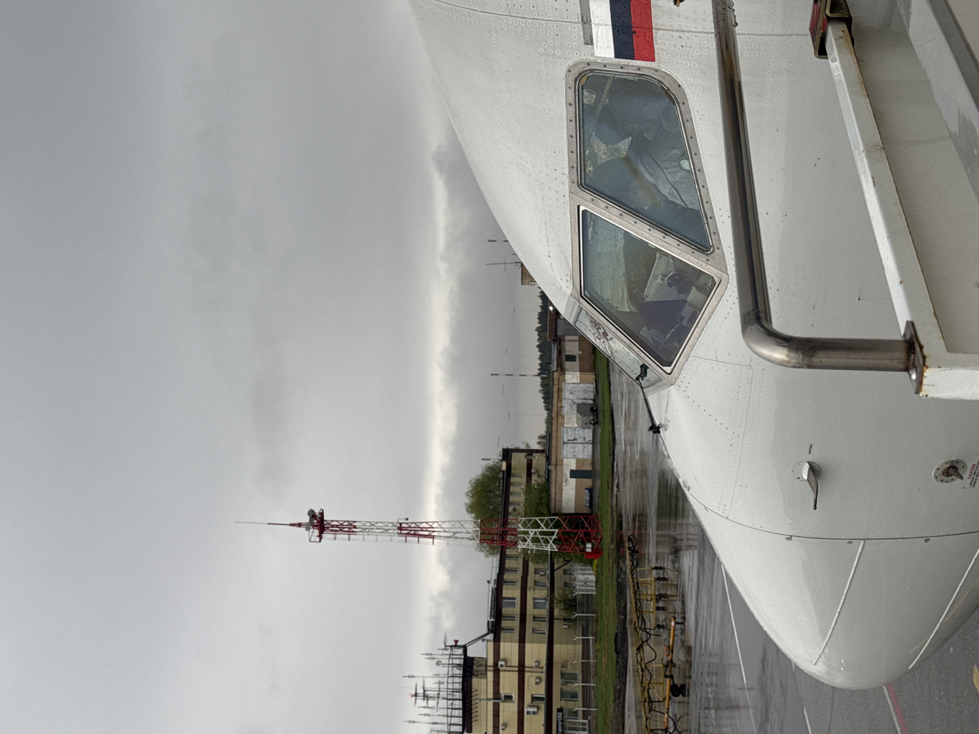 The image depicts the cockpit of an airplane, presumably located at the Bolshoye Savino Airport in Russia, as this airport is close to the city of Bolshoye Savino. The cockpit windows reflect the overcast sky and wet tarmac, suggesting recent rainfall. An antenna tower, painted with red and white sections, stands behind the aircraft. Nearby buildings of a yellowish color are visible, suggesting it may be an airport control center or other terminal building. The immediate environment is characterized by wet, reflective surfaces indicating the presence of rain. There are no people visible in the frame.