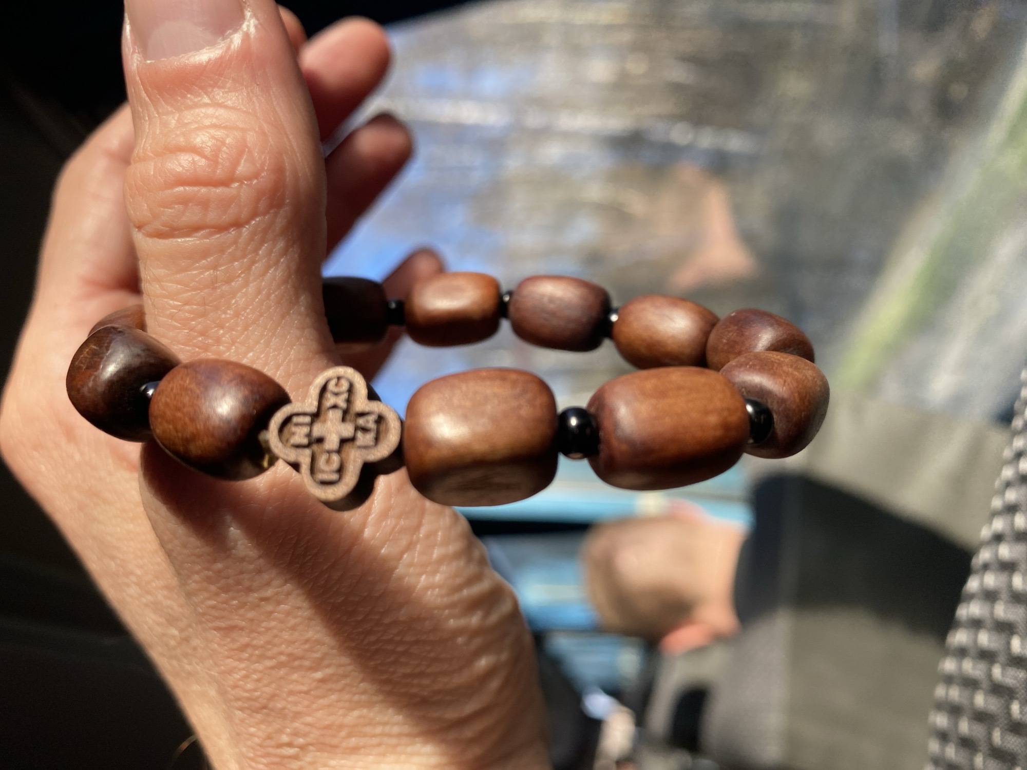 A person's hand holds a wooden bracelet with a cross charm. The bracelet is made of rounded, brown wooden beads, interspersed with small black beads. The cross charm is also made of wood and has a distinct design carved into it. The background shows a blurry interior, possibly a vehicle, with glimpses of someone's head and clothing. The location is not verifiable as Mishutino, Russia based solely on the image.