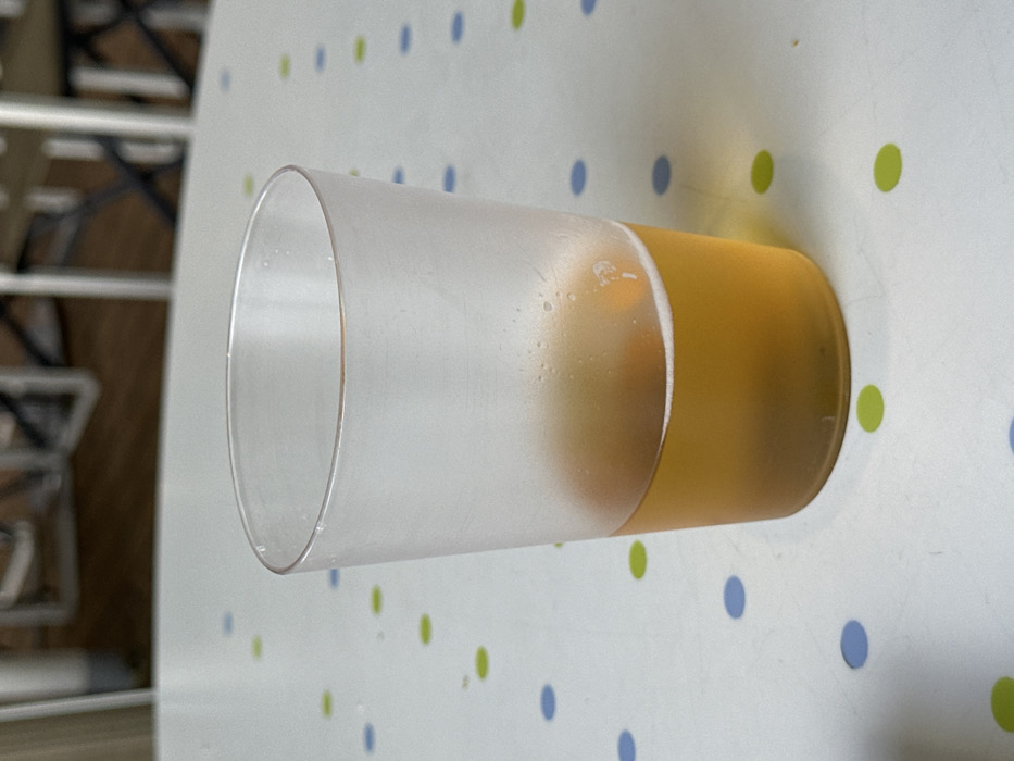 The scene depicts a single translucent plastic cup positioned centrally on a white table surface in an indoor setting within the city of Puerto del Rosario, Spain. The cup is approximately half-filled with a golden-amber liquid, consistent with beer, exhibiting a minimal amount of white foam at the liquid's surface and condensation on the cup's exterior. The table surface features a repeating pattern of uniformly spaced circular dots in alternating light blue and lime green colors. The background is out of focus, showing blurred, light-colored structural elements, likely parts of chairs or other furniture. No individuals are visible, and no discernible actions or interactions are taking place.