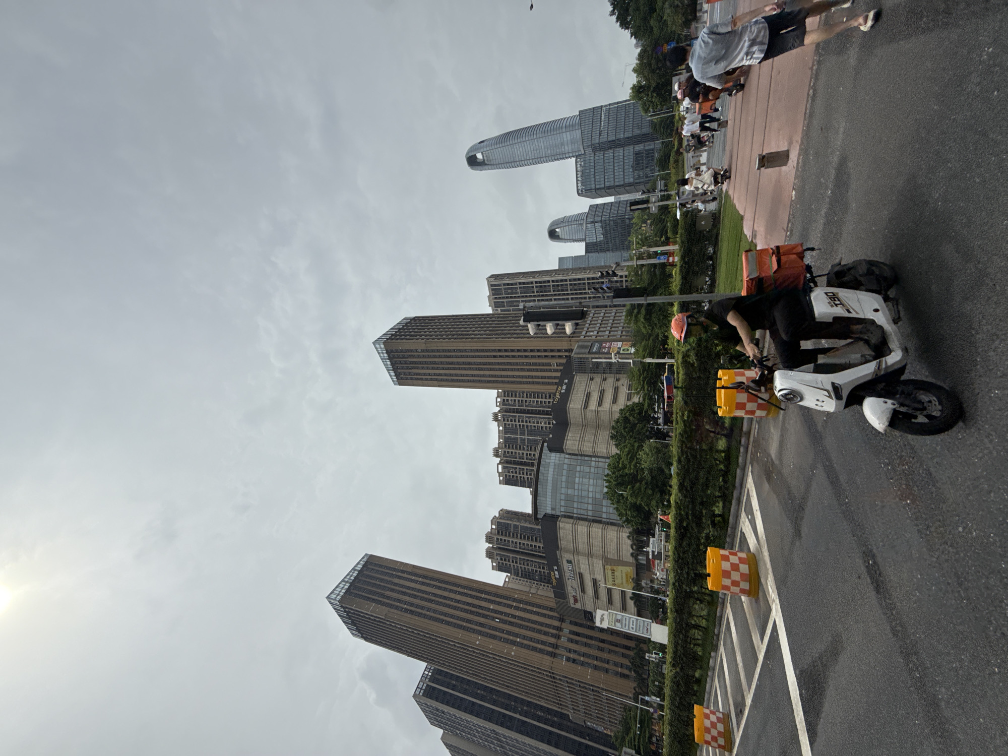 The image depicts a street scene in Haizhu District, China, with a prominent skyline of modern high-rise buildings. The foreground shows a wet asphalt road with traffic markings. A white electric scooter, ridden by a person wearing a black shirt, black pants, and an orange helmet, is in the left half of the frame, facing right. The scooter has a red delivery box mounted on the back. Orange and white checkered traffic barrels are visible on the side of the road. In the middle ground, several pedestrians are seen walking on a reddish pathway. Behind them, a row of lush green trees lines the street. The background is dominated by a cluster of tall buildings, some appearing to be residential or office towers, with distinct architectural features. One building on the far right is a distinctive, curved skyscraper. The sky is overcast and bright, suggesting daytime.