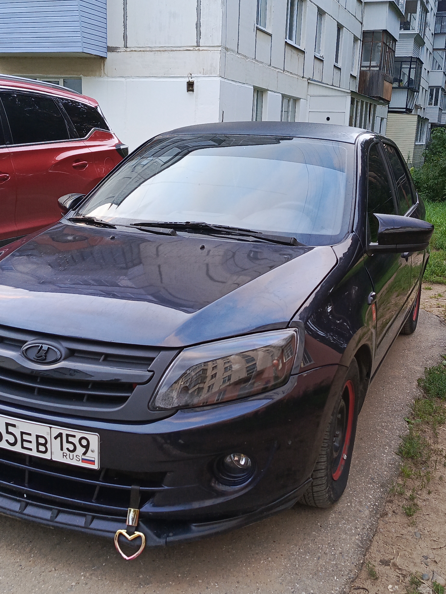 The image shows a dark-colored car parked on a dirt and gravel area next to a sidewalk. The car has a license plate that reads "5EB 159 RUS." A red vehicle is parked to the left of the dark car. In the background, there is a multi-story building with balconies and windows. The scene appears to be in an urban environment. The specific location in the city of Chaikovsky, Russia is not discernible from the image alone.