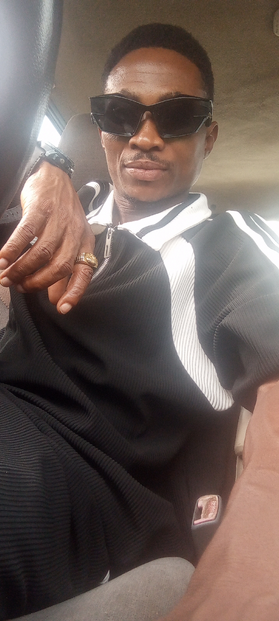 A medium shot shows a dark-skinned man wearing black sunglasses, a black zip-up track jacket with white stripes, and a gold ring. A black wristwatch is also visible on his left wrist. The man is positioned inside a vehicle with gray upholstery. The location, Adankolo, Nigeria, is only mentioned in the prompt.