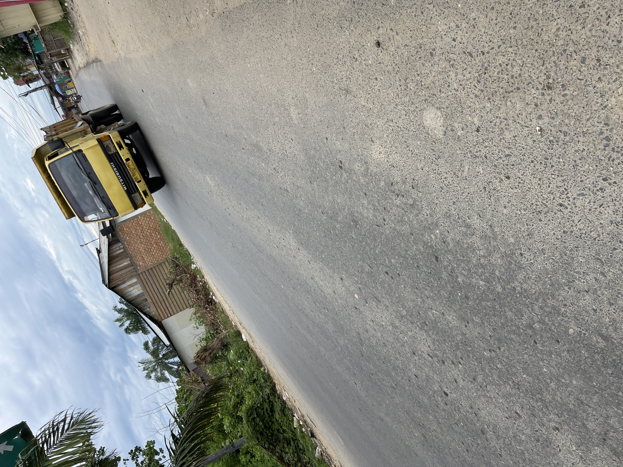 The image shows a bright yellow Mitsubishi dump truck driving on a paved road in Simpangempat, Indonesia. The truck is in motion, with its front wheels turned slightly. The road surface is asphalt, showing signs of wear and tear with scattered small stones and debris. To the left of the road, there is lush green vegetation, palm trees, and some low-lying buildings constructed with corrugated metal and brick. The sky above is partly cloudy. In the background, other buildings and utility poles are visible, suggesting an urban or semi-urban environment. The overall scene depicts a typical day on a road in the region.