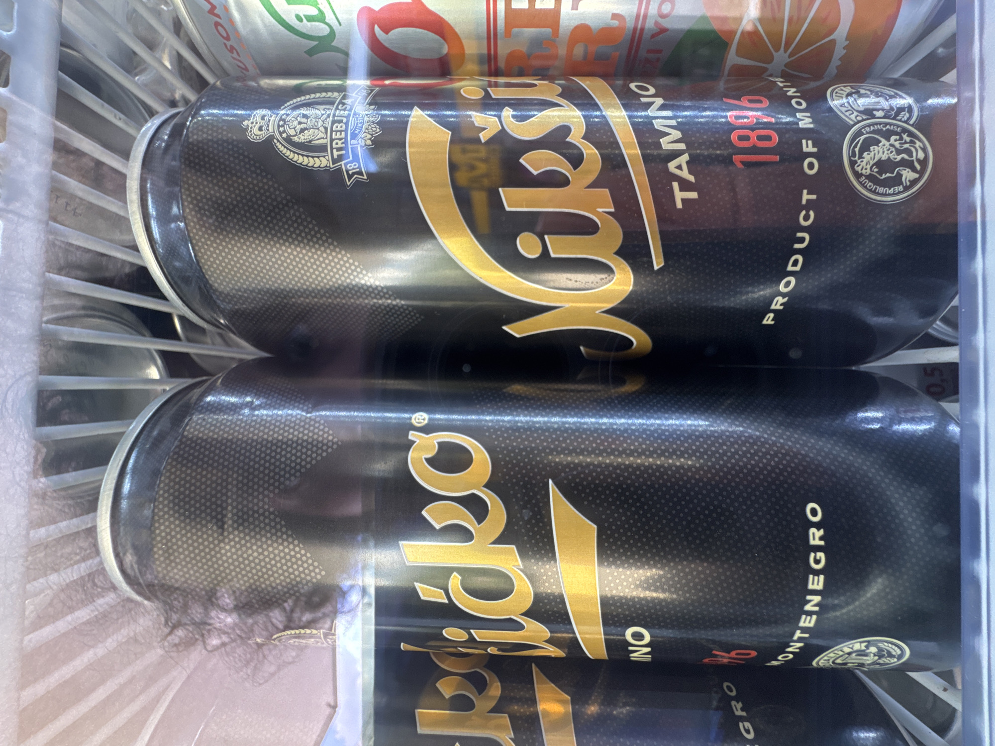 This image depicts the interior of a refrigerated display case in what is identified as a retail setting in Vrijesno, Montenegro. Two black aluminum beverage cans, prominently branded "Nikšićko Tamno," dominate the foreground. Each can features a textured dotted pattern, a gold stylized "Nikšićko" logo, and the word "TAMNO" (dark). The year "1896" is marked in red, accompanied by the text "PRODUCT OF MONTENEGRO." A royal-style crest with "TREBJESA NIKŠIĆ" and "1896" is positioned near the top of the can. At the base of the design, a gold circular medallion inscribed "REPUBLIQUE FRANCAISE" with a profile figure is visible. A third "Nikšićko" can is partially visible on the far left. To the right, a portion of a silver can with an orange graphic and red lettering ("USOM") is discernable. The cans are arranged on a white wire shelf typical of commercial refrigerators. Reflective surfaces show minimal condensation, and strands of dark, curly human hair are visible near the top left portion of the leftmost can, indicating close proximity or interaction.