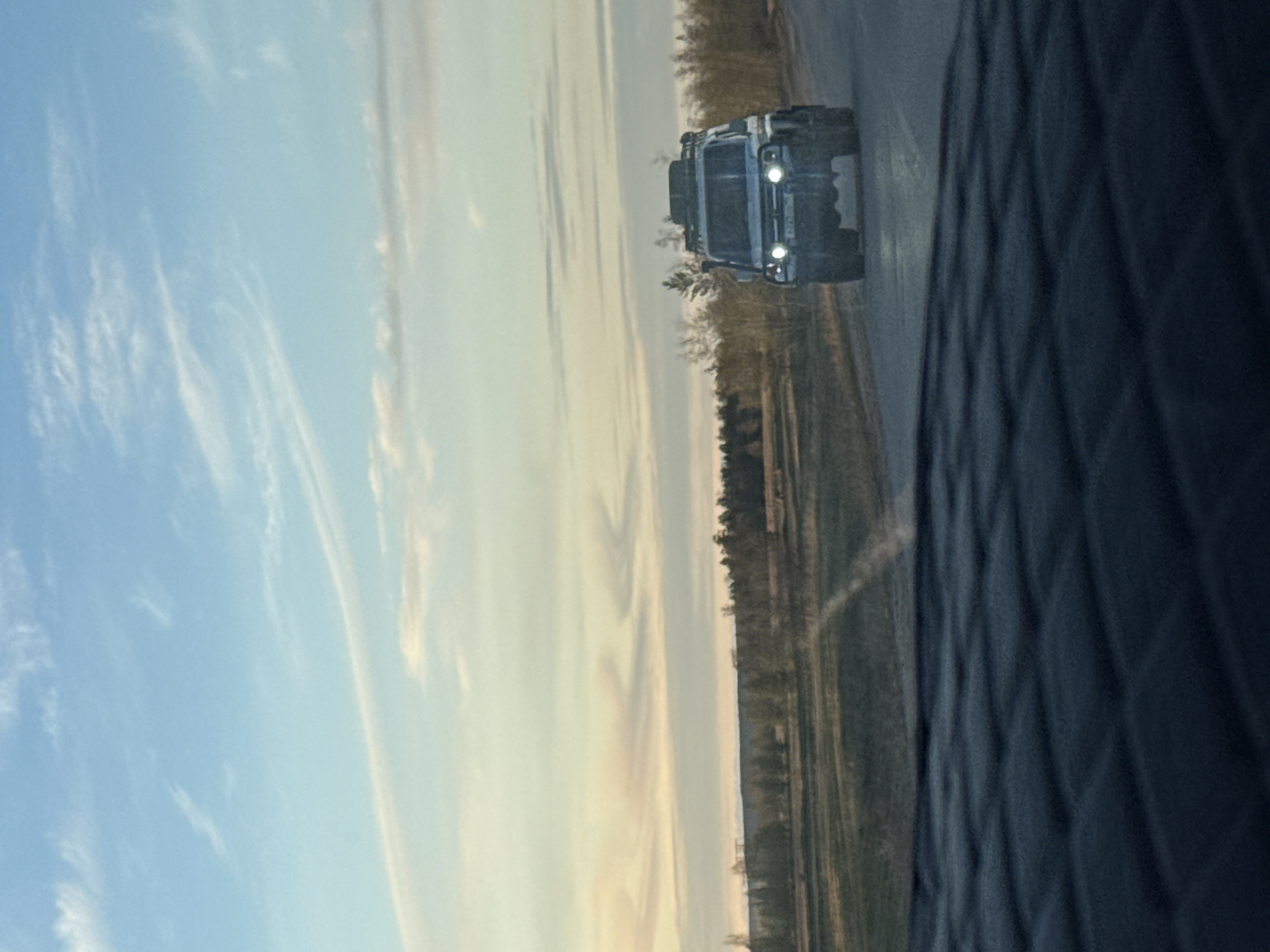 An outdoor, slightly blurry image shows an off-road truck on a road. The image appears to be taken from inside another vehicle, looking through the passenger side window. The truck is light-colored and has a roof rack. Its headlights are on. To the right of the truck is a grassy area with trees. The sky in the background is a mix of blue and yellow, suggesting either sunrise or sunset. Based on the location provided, the scene is in or around the city of Syrdakh, Russia.