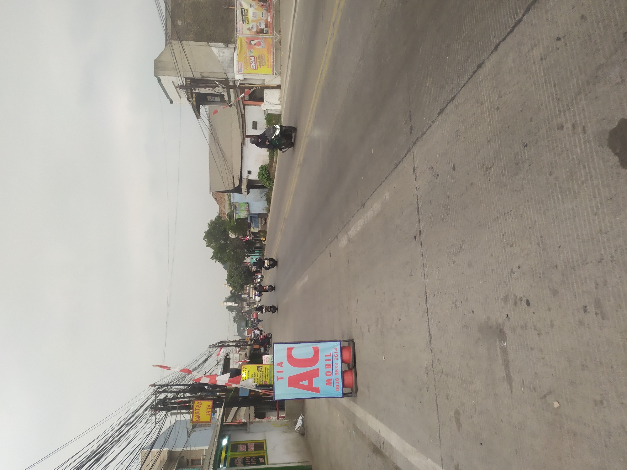 The image depicts a street scene in Kragilan, Indonesia, under an overcast sky. The foreground features a concrete road with faint markings and some scattered debris. On the left side of the road, there is a blue and red sign that reads "TIA AC MOBIL" with a phone number, placed on orange traffic cones. Adjacent to the sign is a building with green trim and a yellow sign that says "WARTEG JAYA." Further down the street, several Indonesian flags are visible on poles.
The road is lined with various buildings, some with visible signage, and a dense network of electrical wires hangs overhead. A few trees are interspersed among the structures. Traffic is present, with several motorcycles and their riders visible on the road. One motorcycle, ridden by a person in dark clothing, is moving away from the camera on the right side of the road. Another motorcycle, also ridden by a person in dark clothing, is traveling in the same direction further ahead. In the distance, more vehicles and buildings are visible, suggesting a commercial or residential area. The overall impression is a typical street in an Indonesian town.